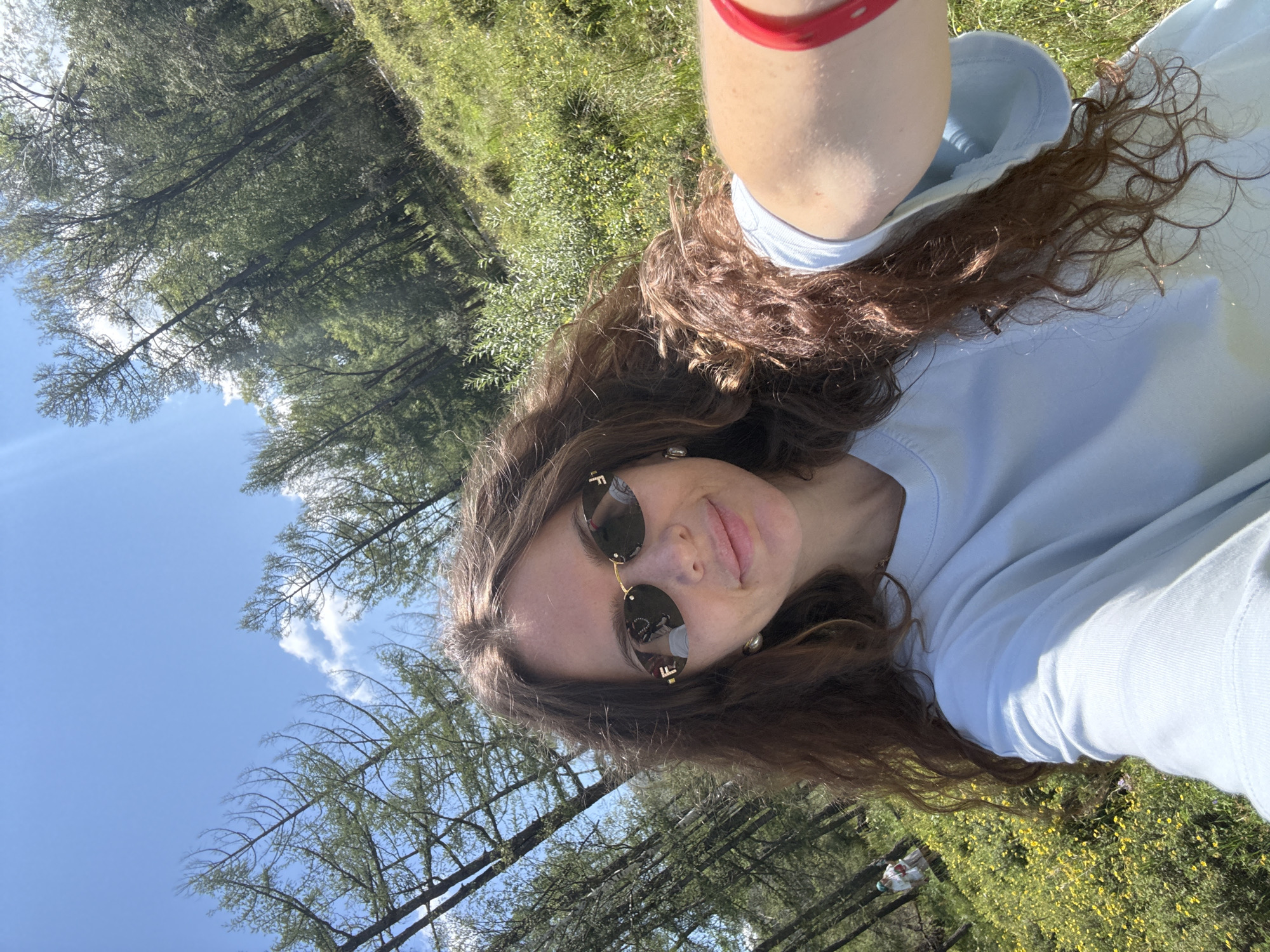 A woman is taking a selfie in a wooded area with a grassy slope covered in small yellow flowers. The woman has long, curly brown hair and is wearing sunglasses, a light blue t-shirt, and a red bracelet on her right wrist. The background shows tall, slender trees, some with sparse needles, and a bright blue sky with scattered clouds. In the distance, on the grassy slope, a person in a white outfit can be seen. The overall scene is bright and appears to be outdoors on a sunny day. The location is identified as Bichiktu-Boom, Russia.