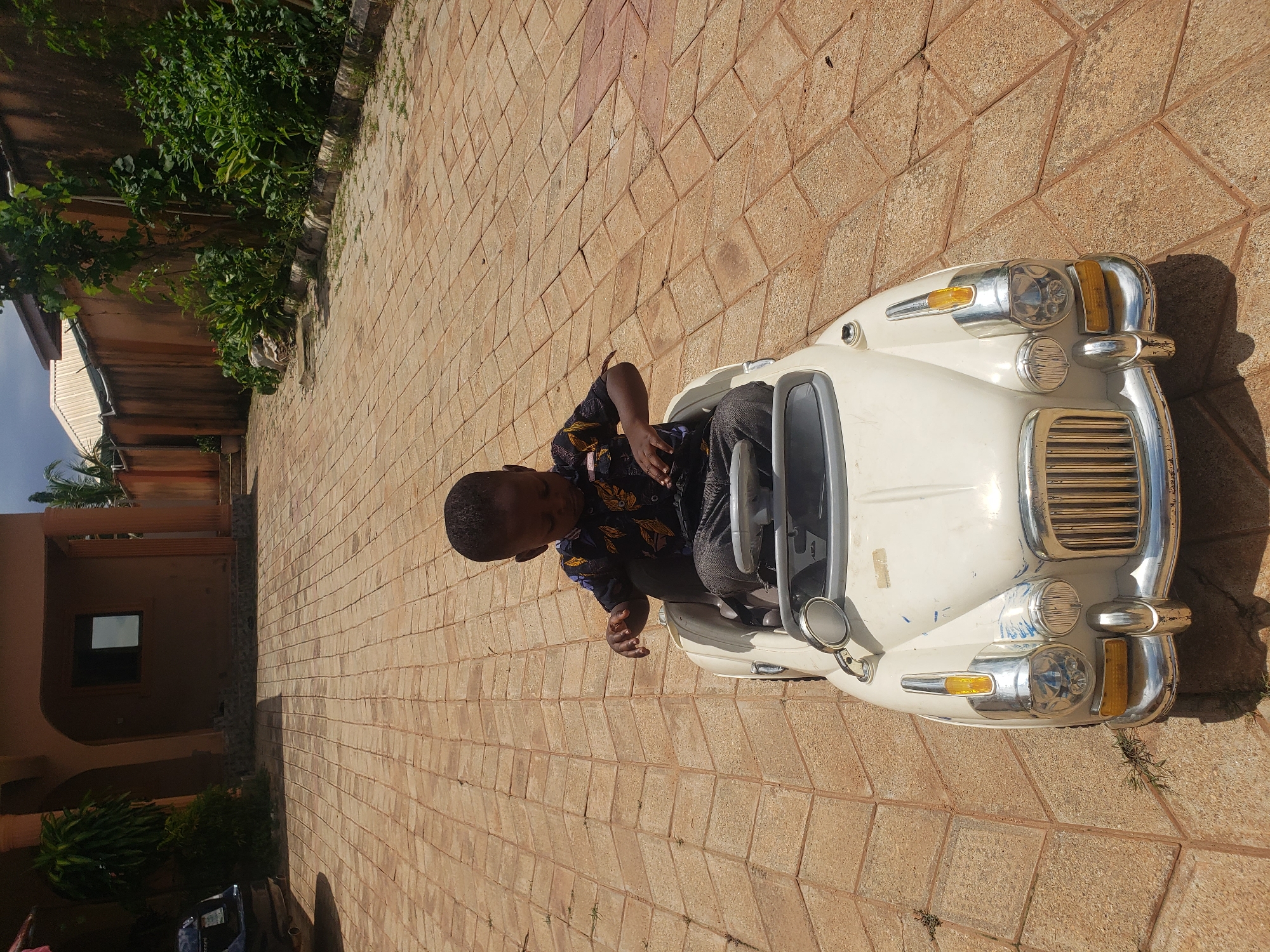 A young Black child sits in a white toy car outdoors on a paved driveway in front of a building. The child is wearing a dark shirt with a floral pattern and dark pants. The car is a classic model with a chrome grille and headlights. The driveway is made of tan pavers arranged in a herringbone pattern. In the background, a light brown building with an arched entryway and a dark window is visible. Lush green vegetation borders the building and a wall in the distance. The scene appears to be set in Sabon Tasha, Nigeria.
