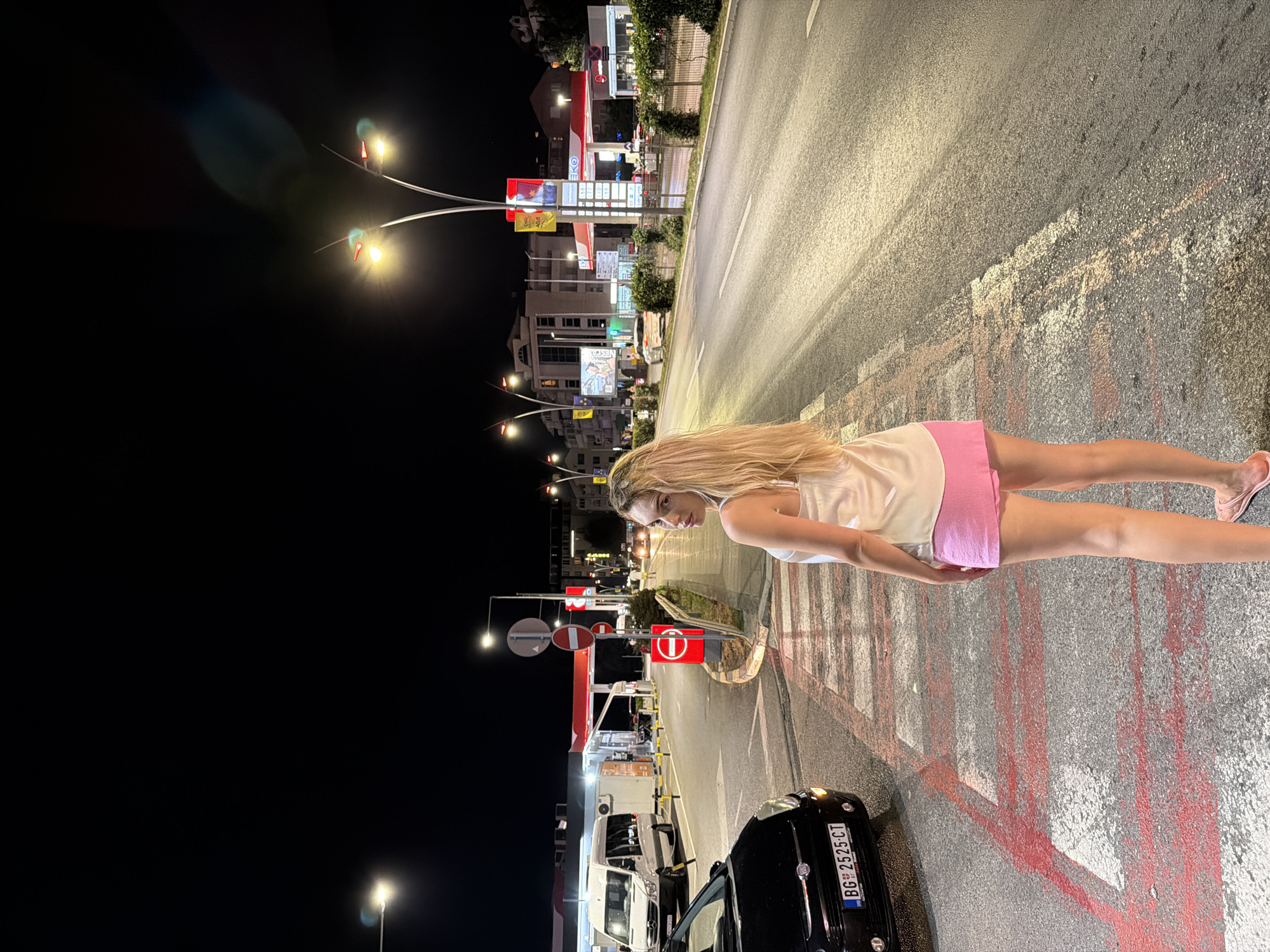 In Budva, Montenegro, a woman with long blonde hair stands on a pedestrian crossing at night. She is wearing a white satin camisole and pink shorts. She is looking over her shoulder at the camera. The street is wet and reflects the lights of the surrounding buildings, streetlights, and a gas station. A black Fiat car is parked on the left side of the pedestrian crossing, and a white van is visible behind it. The street is lined with buildings and shops, and there are several streetlights illuminating the scene. The overall atmosphere is urban and lively, despite the late hour.