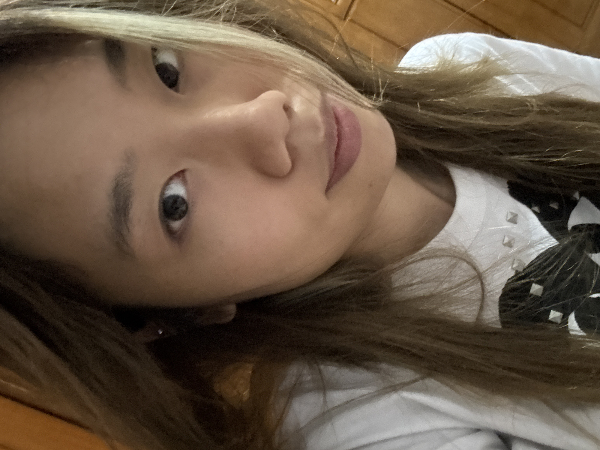 The image shows a close-up, Dutch angle selfie of a young woman lying down. She has blonde-brown hair that falls across her face and shoulders. Her eyes are open and looking towards the camera, and her lips are slightly parted, revealing a hint of pink lipstick. The woman is wearing a white t-shirt adorned with pyramid-shaped studs, some of which are visible on her shoulder. The background appears to be a wooden surface, possibly furniture. The lighting suggests it is daytime. The overall impression is informal and personal. There is no visible text in the image.