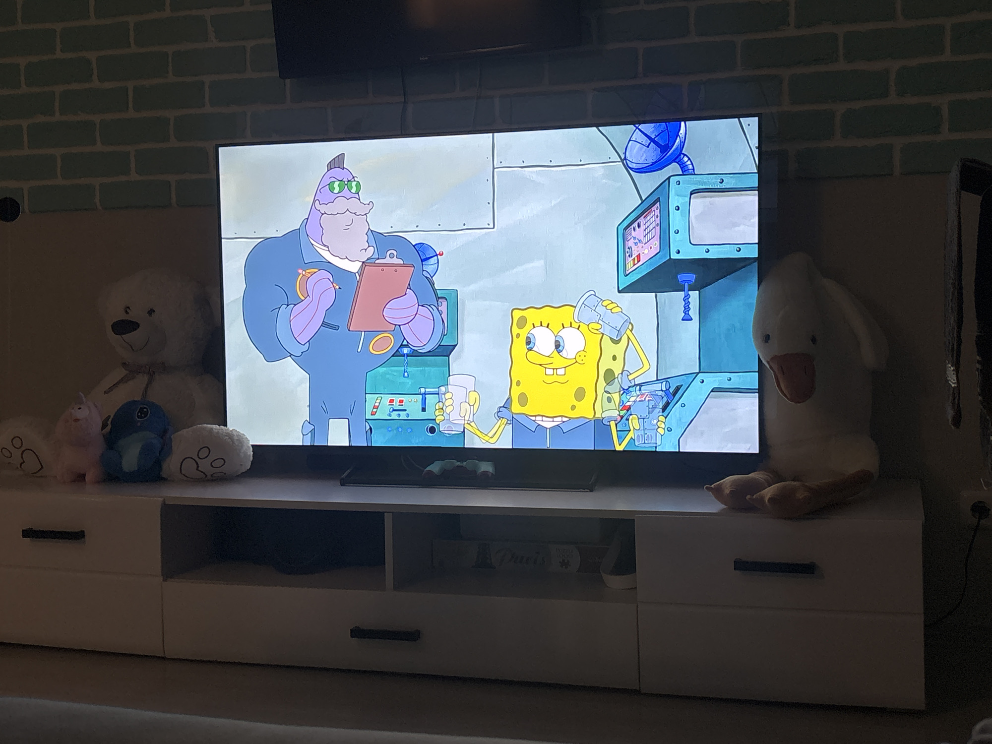The image shows a television displaying a cartoon featuring SpongeBob SquarePants and another character. The TV is placed on a white media console with drawers and open shelves. To the left of the TV are a teddy bear, a pink unicorn, and a blue plush toy. To the right of the TV is a large white stuffed duck. The backdrop consists of a wall made of light green bricks, with a dark object (possibly a speaker) mounted at the top center. The scene is indoors and appears to be in a domestic setting. There is no information to conclude that the location is in the city of Saint Petersburg, Russia.