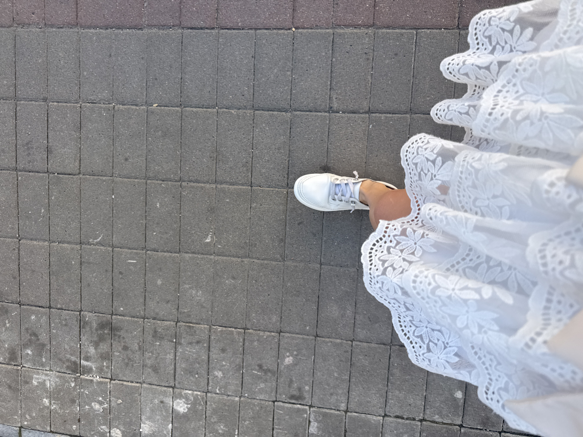 A person wearing a white lace dress and white sneakers is walking on a paved sidewalk. The sidewalk is made of gray rectangular cobblestones. The image is taken from a high angle, looking down at the person's feet and the ground. The person is partially visible, with their left leg and foot in focus. The white lace of the dress is detailed and appears to be a sheer fabric. The context of Saint Petersburg, Russia is not determinable from the image itself.