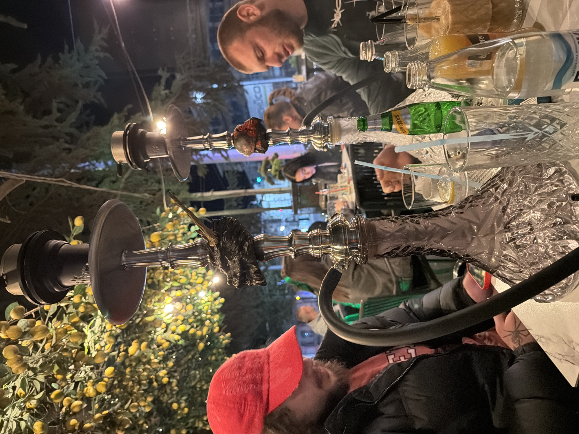 A nighttime scene shows people at an outdoor café, with shisha hookahs in the foreground. A man with a beard and wearing a red cap is looking down at a table. The second hookah has a decorative animal head. Behind the hookahs, there are multiple people sitting at tables, drinking from glasses. Several bottles are also on the table. The location is in a city, as indicated by distant lights and buildings, but the specific city is unknown. Trees and decorative lights surround the outdoor seating area.