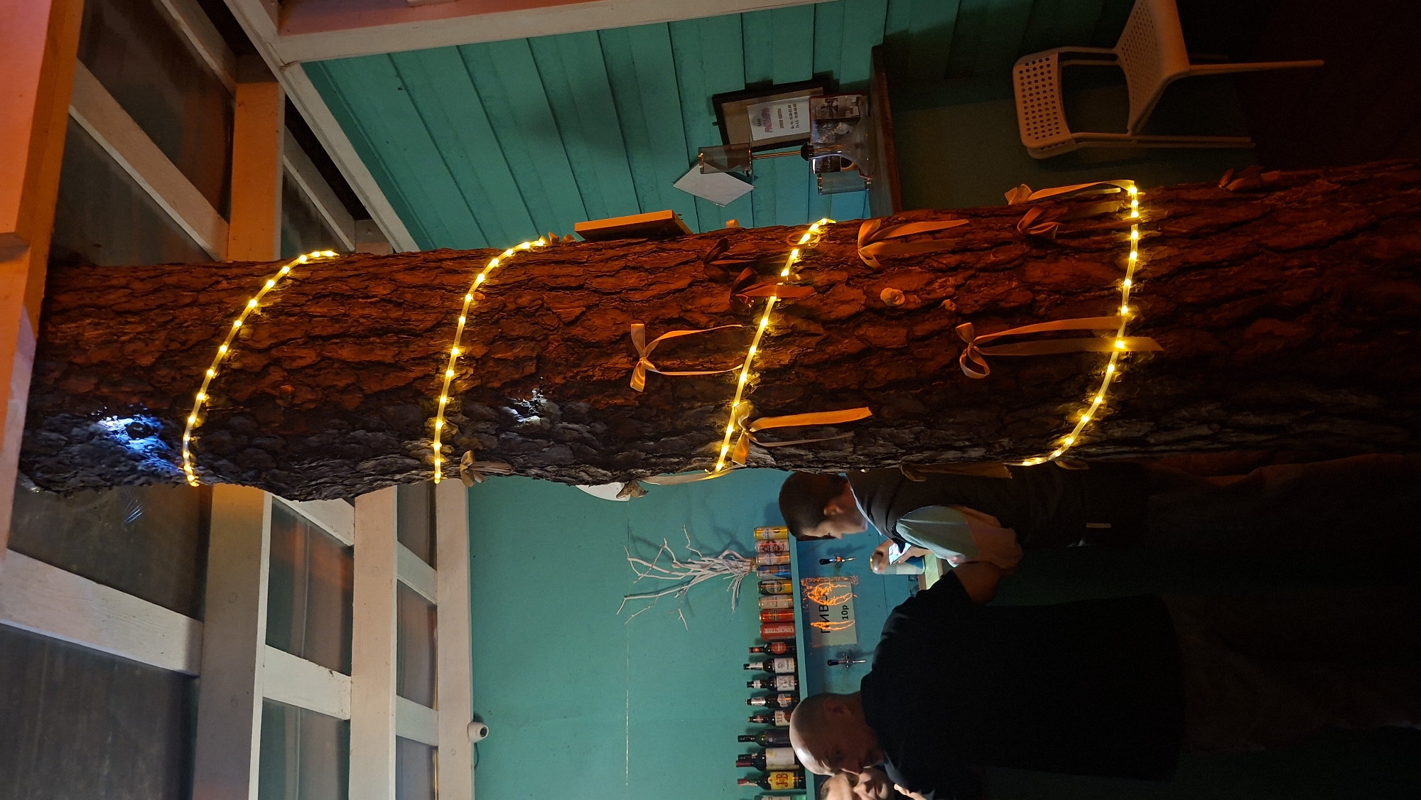 This image depicts an indoor scene, possibly a bar or restaurant, with a prominent tree trunk wrapped in yellow string lights and decorated with orange ribbons. The tree trunk is centrally located and takes up a significant portion of the frame. To the left, two men are visible near a bar counter. One man, wearing a black shirt, is facing away from the camera and appears to be engaged in conversation. Another man, wearing a vest over a lighter colored shirt, is looking at his phone. Behind them, a turquoise-colored wall is visible with shelves holding various bottles and cans. A bare-branched decoration is also present on the shelf. To the right of the tree, a white plastic chair is visible. The ceiling features exposed beams and some lighting fixtures. A security camera is mounted on the ceiling. The overall ambiance suggests a casual, possibly festive, indoor gathering space. The image does not provide specific information about the city of Нарацкі сельскі Савет, Belarus.