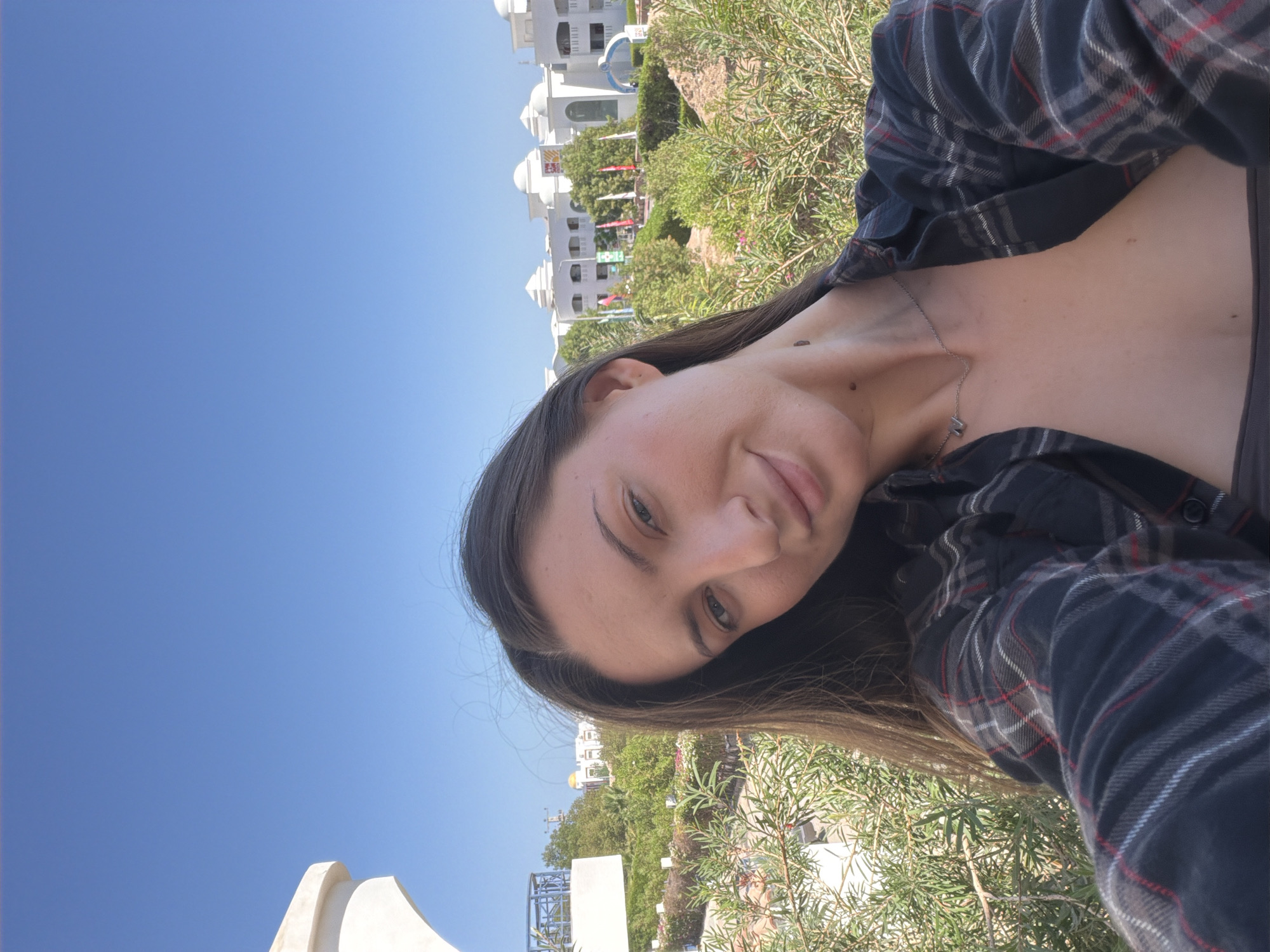 A young woman with long brown hair is captured in a selfie taken outdoors under a clear blue sky. She is wearing a dark plaid shirt with red and white accents. The image is taken from a high angle, with the woman's face tilted towards the camera. She appears to be smiling or about to smile, with a relaxed and pleasant expression. A delicate silver necklace is visible around her neck, and a small mole is noticeable on her collarbone.
In the background, a cluster of white buildings with rounded domes and arched windows suggests a resort or hotel setting. Green foliage and some flowering plants are interspersed between the buildings and in the foreground, indicating a warm climate. The bright sunlight suggests it is daytime, likely mid-morning or afternoon. The overall impression is one of leisure and enjoyment in a sunny, possibly tropical or Mediterranean, location. Although not explicitly stated, the context provided places this scene in Sharm El Sheikh, Egypt.