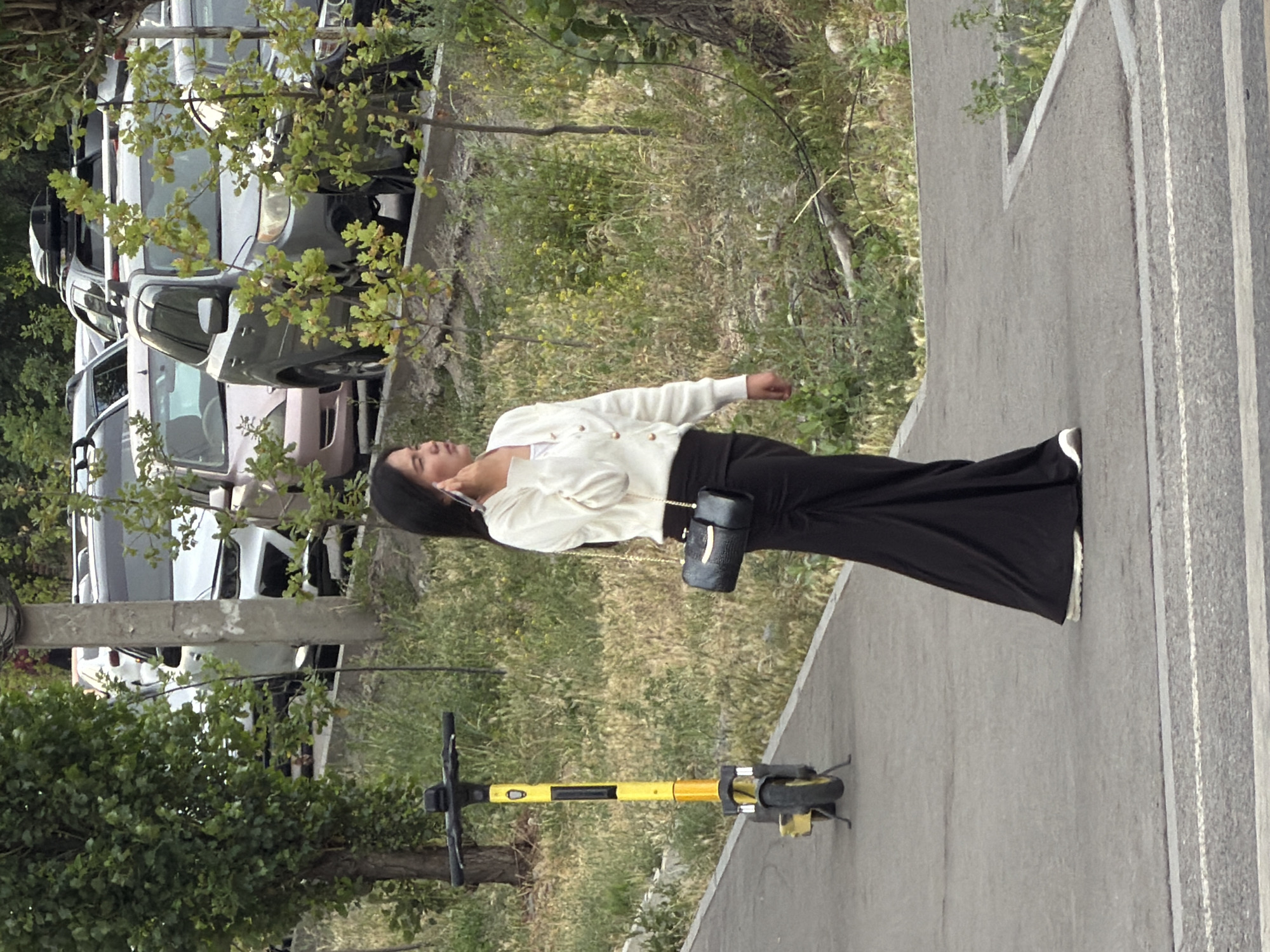 A woman with dark hair is standing on a paved walkway in an urban outdoor setting, talking on her cell phone. She is wearing a white long-sleeved cardigan over a white top, a long black skirt, and white sneakers. She is carrying a small, dark, textured shoulder bag with a gold chain strap. To her left, a yellow electric scooter with black accents is parked on the walkway. Behind the woman, a line of parked cars is visible, including a silver SUV and a light pink sedan. The area is characterized by green grass, scattered trees, and some smaller vegetation. The background shows more trees and a pole with wires.