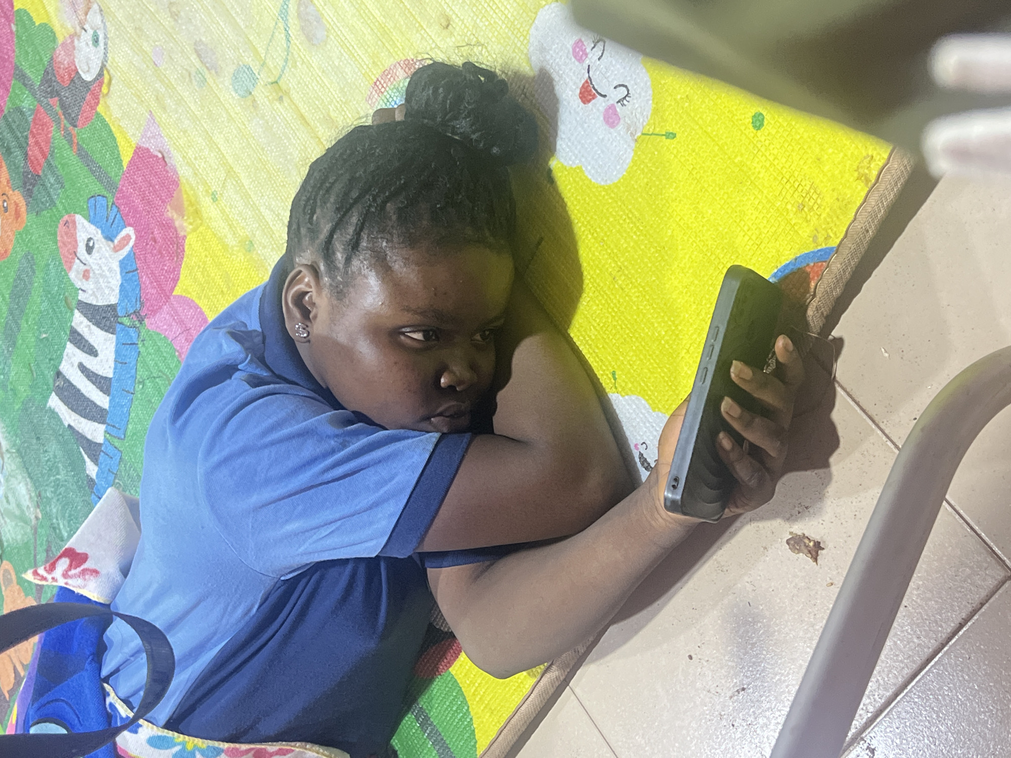 A person is depicted lying on a colorful play mat indoors, looking at a smartphone. The subject, likely female, has dark skin and braided hair tied in a bun. She is wearing a light blue short-sleeved shirt with a darker blue collar and sleeve cuffs, possibly a uniform or polo shirt. Her head is resting on her left arm, while she holds a dark-cased smartphone with both hands, focusing intently on its screen. Her expression appears serious or contemplative.
The play mat, where she is resting, is vibrantly colored with sections of yellow, green, pink, and white, featuring cartoon illustrations including a zebra, a smiling cloud, and musical notes. Part of a blue bag with a colorful, patterned strap is visible in the bottom-left corner of the image. Adjacent to the mat is a light-colored tiled floor, on which a curved, light grey metal bar (possibly part of furniture) is situated. Some small, dark specks, resembling debris or food crumbs, are scattered on the tiled surface. The scene suggests a relaxed, indoor environment, possibly a home in Obosi, Nigeria. The lighting is soft and artificial. No explicit text is visible in the image.
