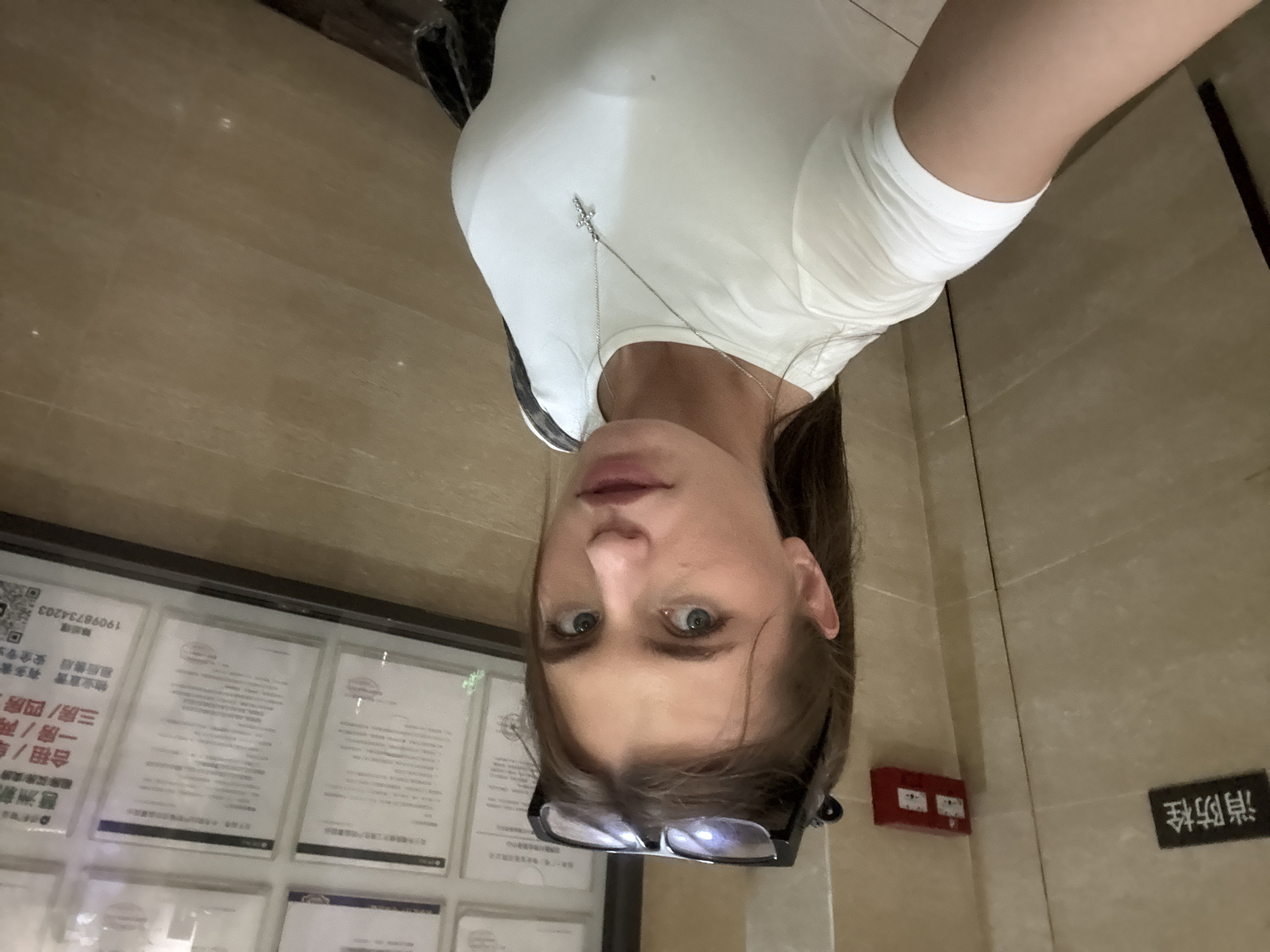 A young woman with blue eyes and brown hair, wearing a white t-shirt and a silver cross necklace, is taking a selfie in an elevator. She has glasses perched on top of her head. The elevator walls are tiled in a light brown, wood-grain pattern, and there are several framed documents on the wall. A red fire alarm box is visible on the left wall, and a sign with Chinese characters, "消防栓" (fire hydrant), is also present. The floor of the elevator appears to be a light brown laminate.