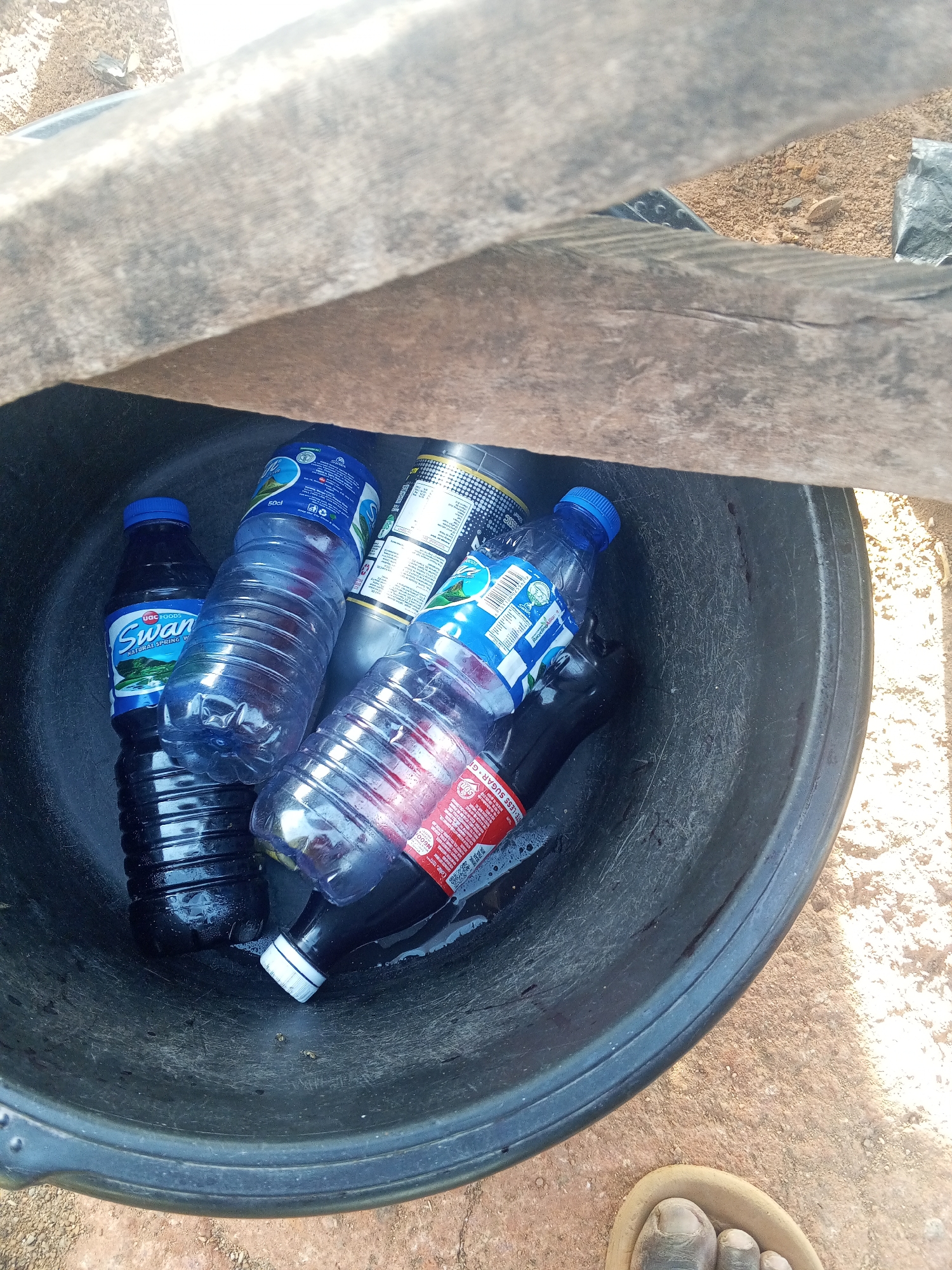 Gwong, Nigeria: A black bucket contains several discarded plastic bottles (including "Swan" and "Coca-Cola" brands) and a metal can. A wooden plank rests partially over the bucket. A bare foot wearing a sandal is visible in the lower right corner. The bucket is on a reddish-brown surface.