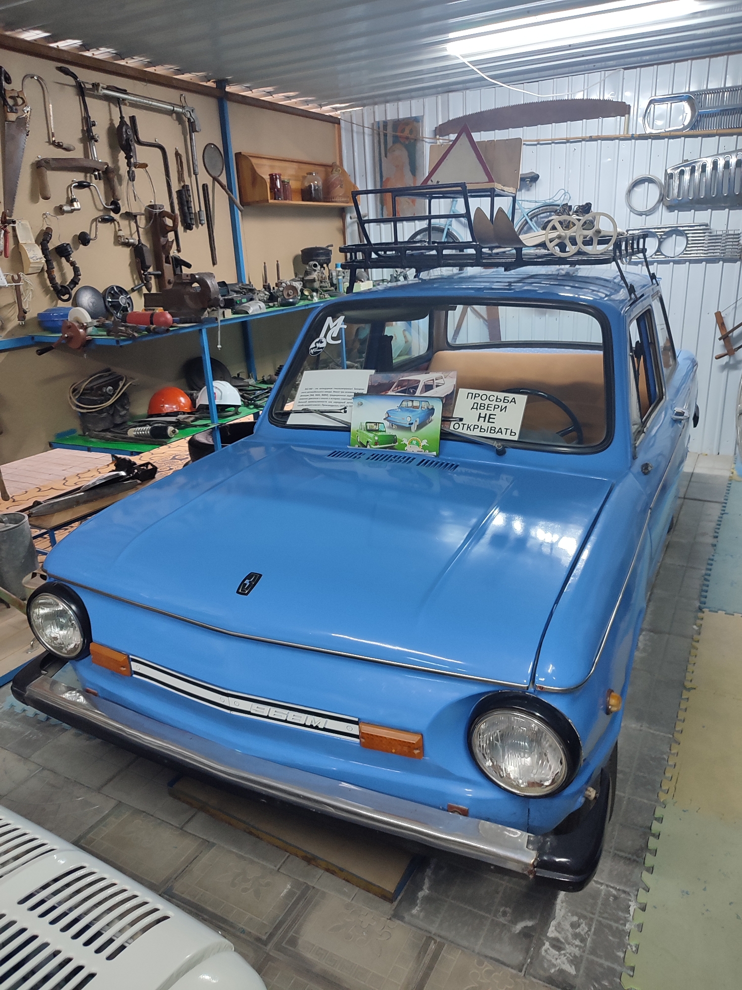 A light blue, vintage car is parked inside a garage. The car, a ZAZ 968M, has a luggage rack on its roof. On the dashboard, a sign reads "PROSIM, DVERI NE OTKRIVAT", which translates to "PLEASE, DO NOT OPEN THE DOORS". The garage walls are lined with shelves displaying various tools and car parts, suggesting it might be a workshop or a museum. The floor is covered with tiled mats. The car is the primary subject of the image, with the surrounding environment serving as context. The city of Qonaev, Kazakhstan, is mentioned as the location, which might indicate a local museum or a private collection. There are no people visible in the image, and the car appears to be stationary.
