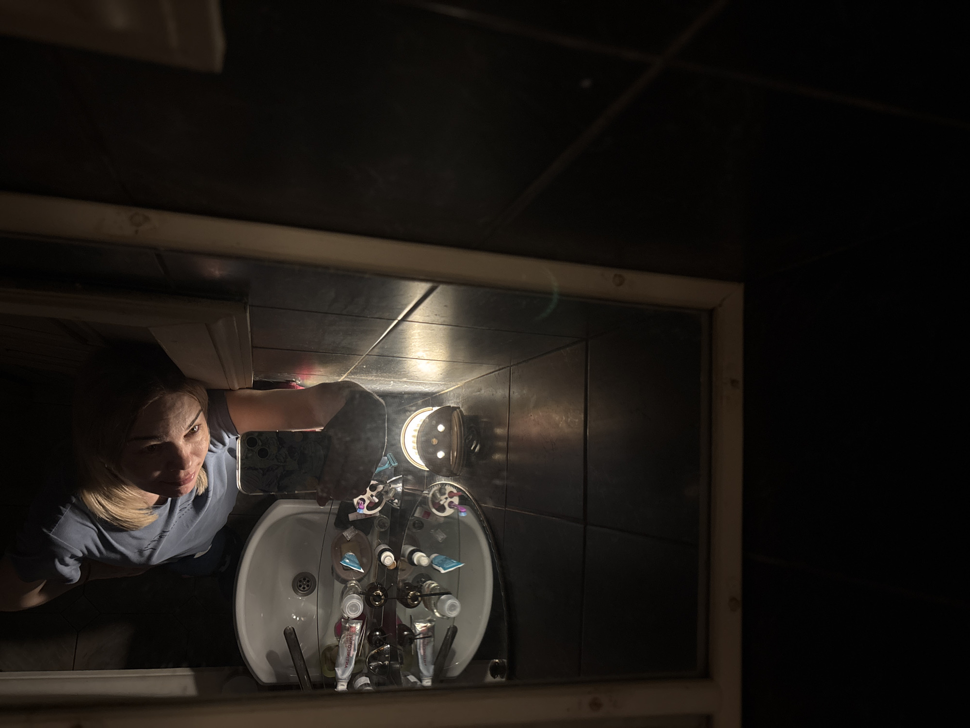 A selfie is taken in a darkly lit bathroom in Vsevolozhsk, Russia, as reflected in a mirror. A woman with blonde hair and a light blue shirt holds a smartphone, partially obscuring her face in the mirror’s reflection. Below the mirror, a white porcelain sink is mounted on a dark tiled wall. Several toiletries, including tubes and bottles, are arranged on the sink’s surface. A light fixture above the sink illuminates the area. Dark tiles cover the wall and floor, and the rest of the environment is obscured in darkness.