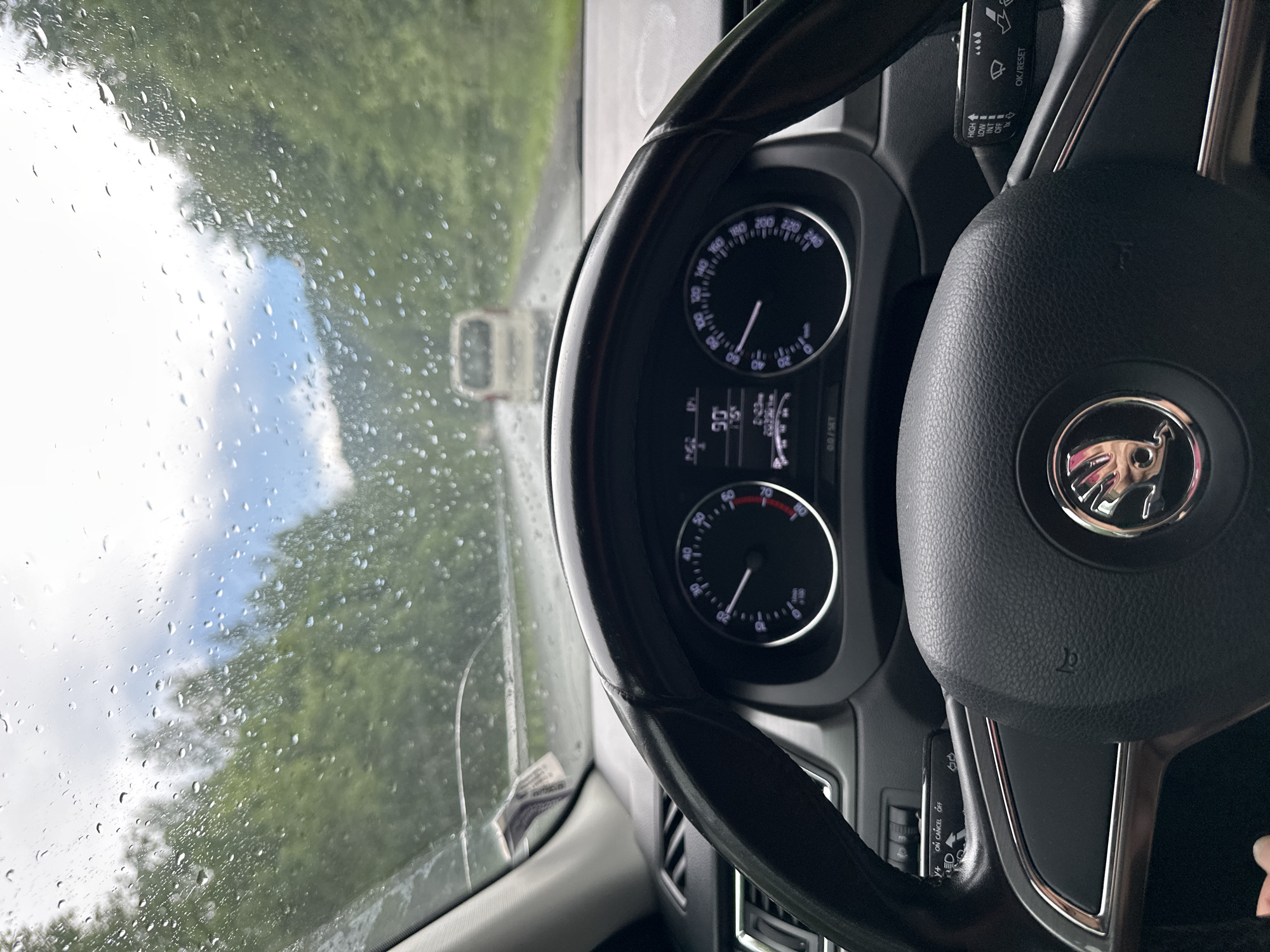 The image captures the interior of a car, looking out through a rain-streaked windshield. The driver's perspective is evident, with the steering wheel prominently displayed in the foreground. The dashboard features two main circular gauges, likely a speedometer and a tachometer, with a smaller digital display between them showing "90" and other unreadable data. The steering wheel has a Skoda emblem in the center. To the right of the steering wheel, a turn signal stalk is visible. Outside the car, the road is wet, and trees line both sides, suggesting a rural or forested area. A white van is visible ahead on the road, and the sky appears overcast with patches of blue. The scene conveys a sense of driving in the rain. The location is specified as Brishbash, Russia.