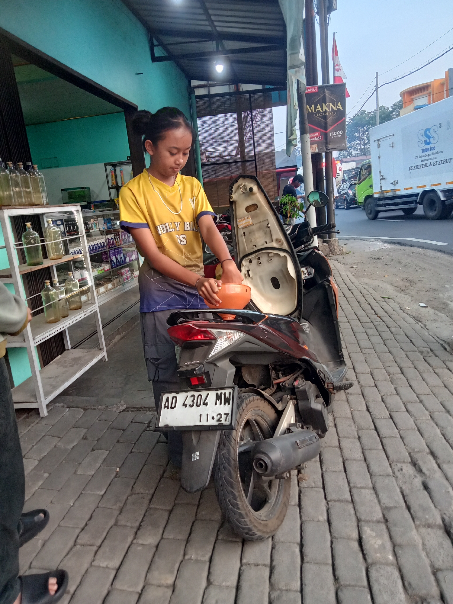 A young female is standing by a dark-colored motorcycle with its seat open. She is wearing a yellow t-shirt with blue and purple accents and gray cargo pants. She is holding an orange funnel or container over the open seat area, possibly to pour something into the motorcycle. The motorcycle has a license plate that reads "AD 4304 MW" with "11-27" below it.
The scene appears to be on a street in Tegalrejo, Indonesia, indicated by the presence of a nearby shop with shelves stocked with bottles and various items. The shop has a turquoise-colored exterior wall. A white shelf unit filled with bottles is to the left of the young woman. In the background, there is a road with traffic, including a white truck with the text "SSS Tube Ice" and a green truck. A banner with the text "BARU MAKNA EXCLUSIVE" is hanging from a pole. The ground is paved with gray interlocking bricks. The lighting suggests it is daytime, with a clear sky.