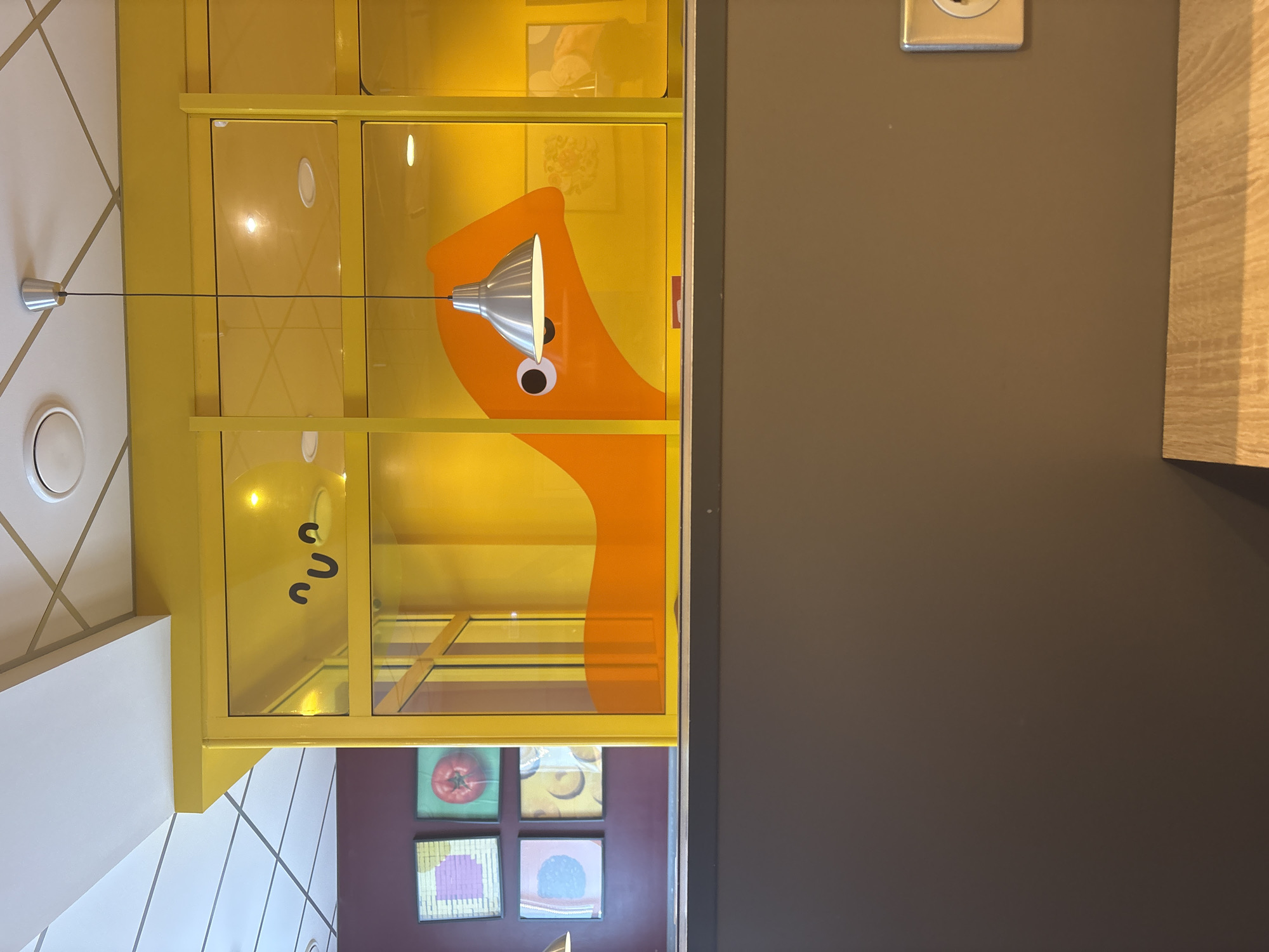 The image shows an interior scene, possibly of a business or restaurant. Visible are a yellow cabinet with glass doors featuring an orange cartoon character on the interior, as well as artwork of vegetables. The environment includes light-colored geometric tile design visible on the walls and ceilings. The scene also captures a portion of a dark gray wall and a light-colored wooden panel. A light fixture hangs in front of the cabinet. Based on the artwork in the corner, it is likely the location is in the city of Votkinsk, Russia.