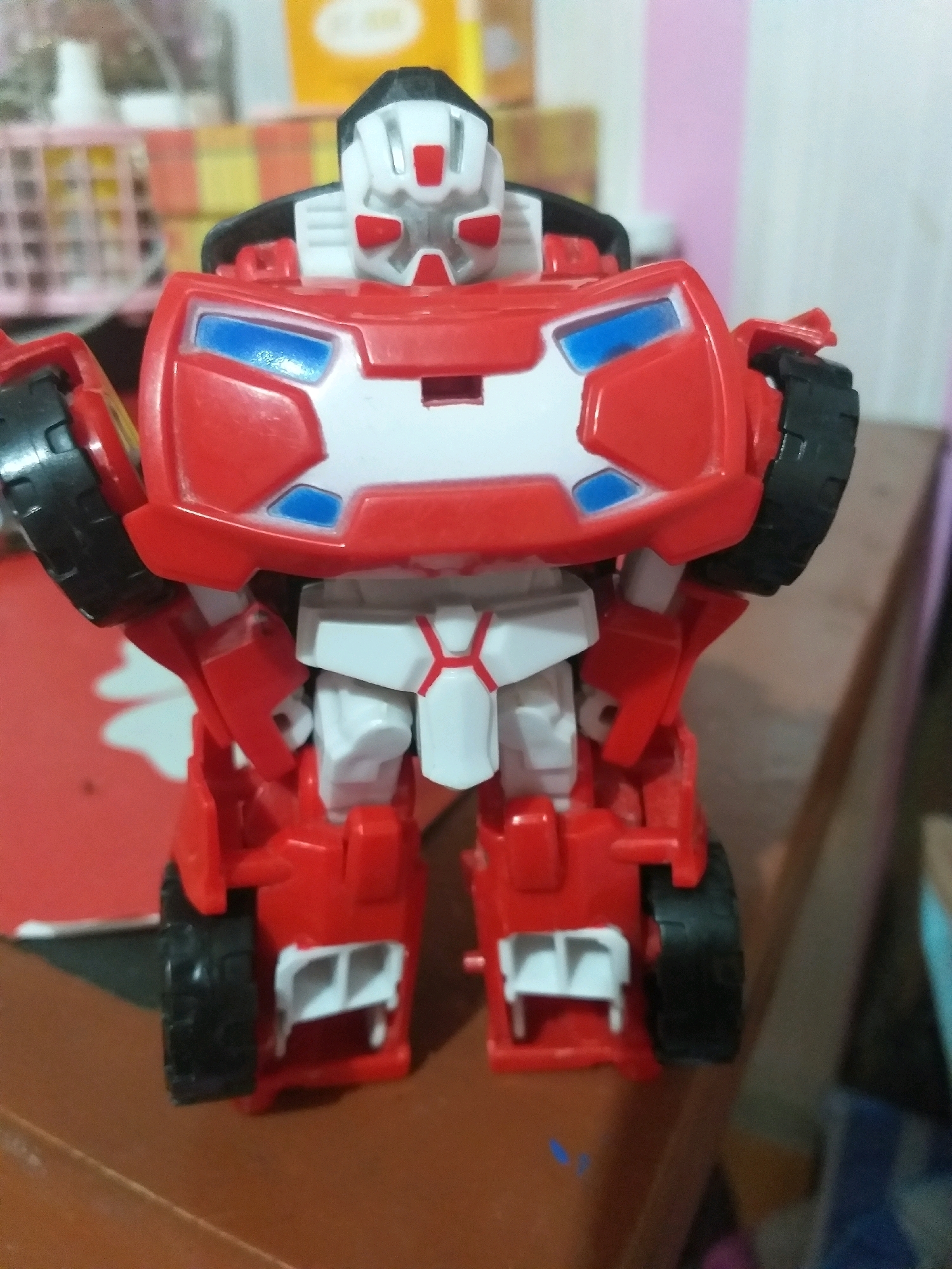 The image displays a close-up, eye-level, vertical shot of a red and white toy robot. The robot is standing on a brown wooden surface. The robot is primarily red with white accents and blue detailing on its chest. It has black wheels on its sides, suggesting it can transform into a vehicle. The background is blurred and appears to be an indoor setting with shelves and objects that are out of focus. The lighting suggests an indoor environment, possibly a room or a store display. There is no discernible indication of the city of Dumai City, Indonesia, within the image itself, as it is a general indoor scene with a toy.