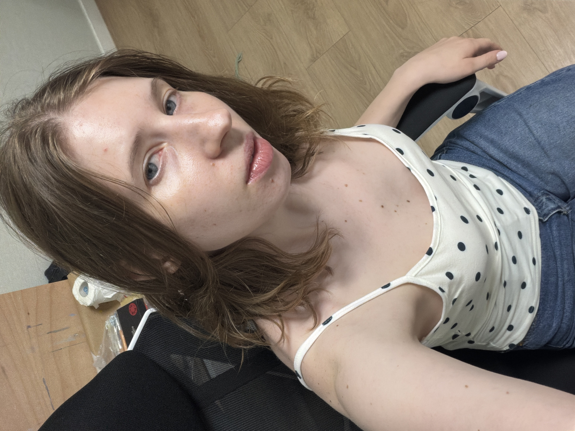 The image shows a young Caucasian woman with light brown hair and blue eyes sitting in a black office chair. She is wearing a white polka dot tank top and blue jeans. Her head is tilted and she is looking slightly off-camera. The background appears to be an indoor setting, with a wooden floor and a plain white wall. There are some items visible in the background, including a roll of toilet paper and some packaging. There is no visible indication of the location being in Dongducheon-si, South Korea.
