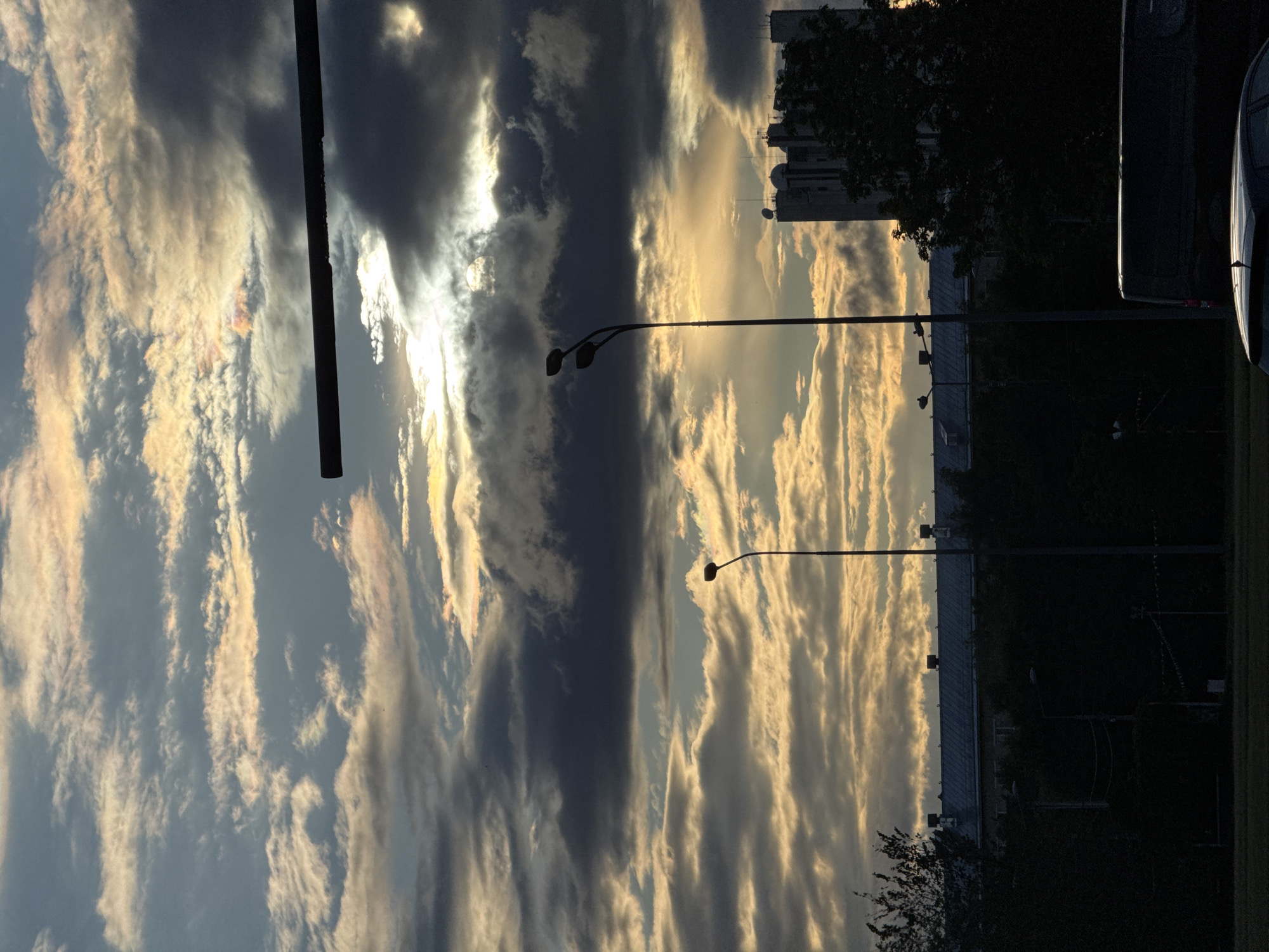 This is a vertical, eye-level, medium shot of a city skyline against a dramatic, cloudy sky at dusk. The sky is filled with horizontal bands of clouds, ranging from dark gray to pale yellow and pink, with the sun peeking through in the upper center. A black horizontal bar, possibly a traffic signal or part of a bridge, cuts across the upper portion of the frame. In the mid-ground, silhouetted against the bright sky, are two streetlights with multiple bulbs. Further down, a long, low-rise building with a dark roof and visible chimneys is visible, partially obscured by dark trees. To the right of the low-rise building, a tall, modern building with satellite dishes on its roof stands out. In the foreground, silhouetted dark shapes suggest trees, a street, and the tops of vehicles. The scene appears to be in an urban environment during the evening.