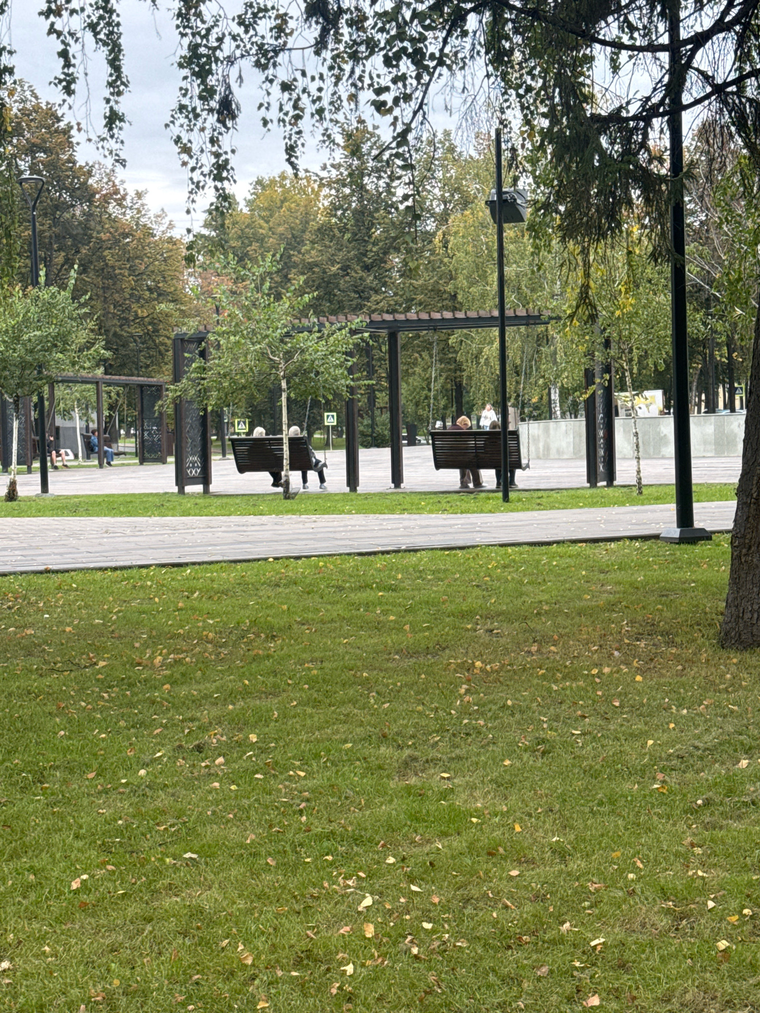 In the city of Ufa, Russia, an outdoor park scene unfolds on an overcast day. In the foreground, a lush green lawn is dotted with fallen yellow leaves, suggesting autumn. A grey paved walkway runs horizontally across the middle ground. Beyond the walkway, a shaded seating area features several wooden swings with built-in benches. On one of these swings, two elderly individuals, a man and a woman, are seated side-by-side, facing away from the camera. The man is wearing a dark jacket and trousers, while the woman is in a light-colored top and dark trousers. Both appear to be conversing or simply enjoying the park. Further back, other swings are visible with other individuals seated on them, including a person in a blue jacket and another in darker clothing. The background consists of dense trees with foliage in various shades of green and yellow, indicating the season. Several lampposts and decorative wooden structures are interspersed throughout the park.