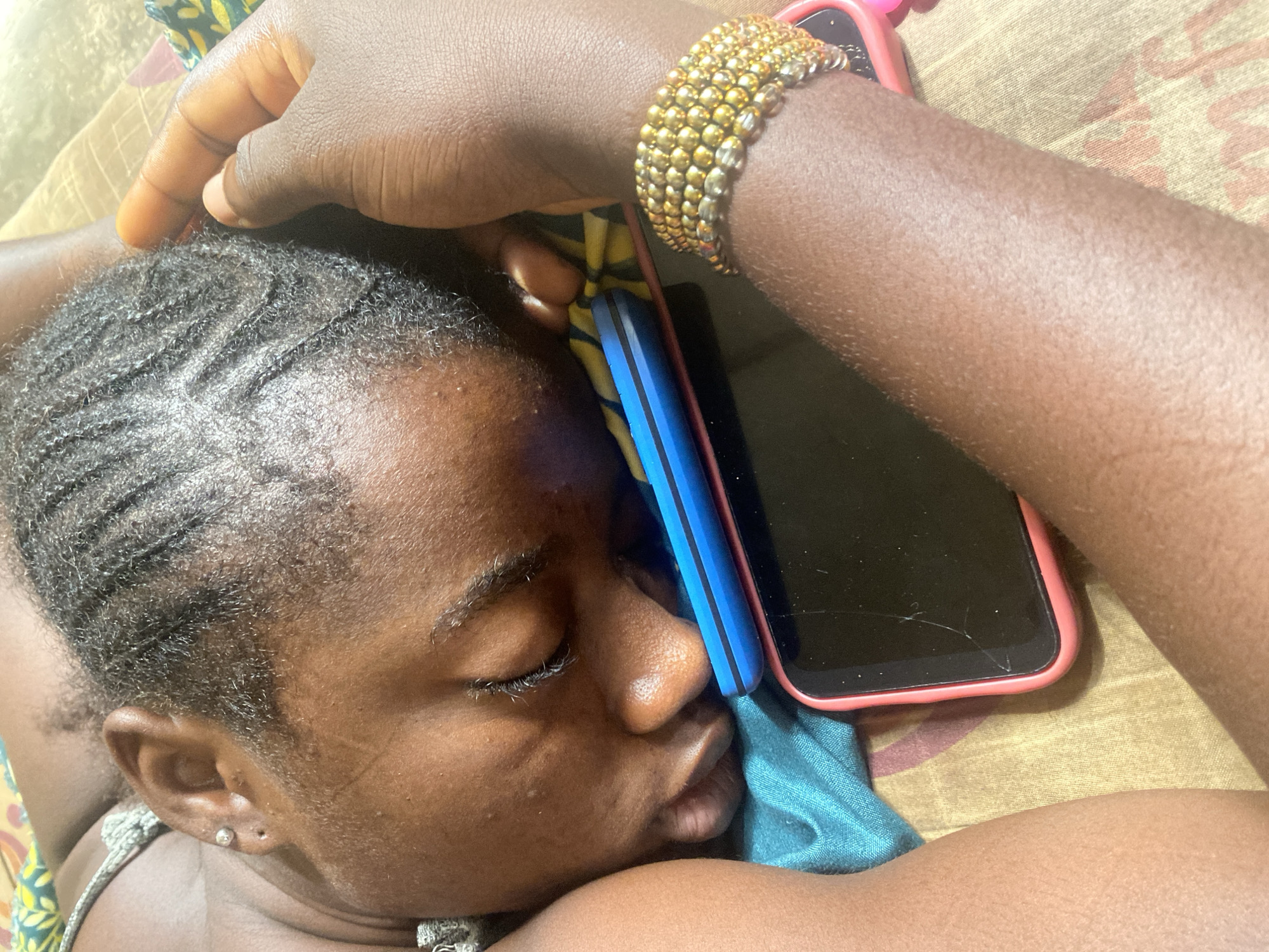 The image captures a close-up, high-angle view of a person, likely female, resting or sleeping in an indoor setting, possibly a bed or couch, in Ikorodu, Nigeria. The individual has a dark skin tone, with eyes closed in a peaceful expression, suggesting sleep or deep rest. Their hair is styled in neat, dark cornrows. A small, silver-toned stud earring is visible in their left ear. There appears to be a faint mark or scar on their left cheek.
Their right arm, adorned with a multi-strand gold-beaded bracelet, is resting near their head. Two smartphones are prominent in the frame: one, with a blue and black design, is nestled close to the person's head, appearing to be used as a pillow or held against the ear. The second phone, encased in a pink or red protective cover, lies beside it, face-down on the fabric, revealing a distinct crack across its screen. The background consists of patterned fabric, possibly bedding or a blanket, with earthy tones and abstract designs. The lighting is soft, indicating an indoor environment without harsh direct light. No visible text is present in the image. The overall scene conveys a moment of quiet rest.