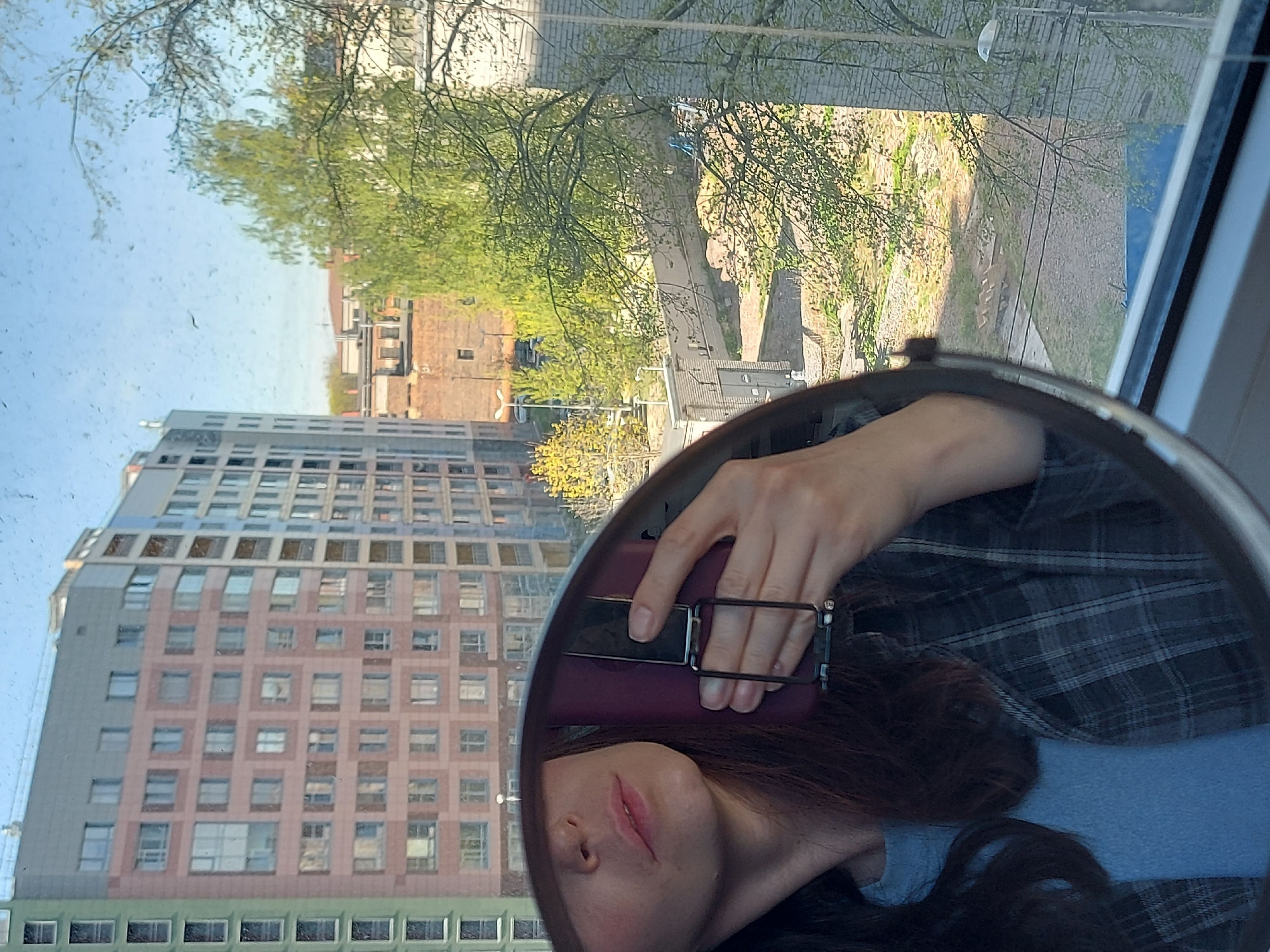 The image shows a reflection in a mirror. A person is holding a smartphone, partially obscuring their face. The mirror's reflection reveals an urban scene with a multi-story building with a pink and gray facade and windows arranged in a grid-like pattern. Other buildings are visible in the background, along with trees. A window frame surrounds the mirror and the reflected scene. The location is suggested to be Saint Petersburg, Russia, based on architectural styles.