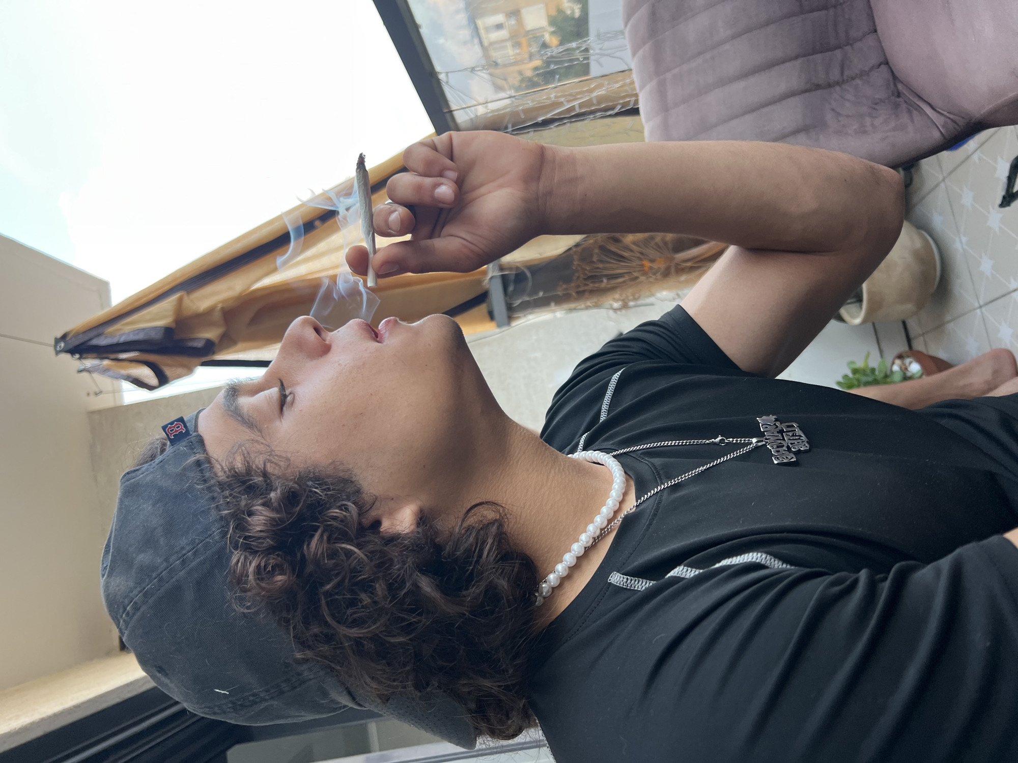 A young man with curly brown hair, wearing a black baseball cap and a black t-shirt with a silver chain necklace, is depicted from the chest up, in profile, on a balcony. He is holding a lit cigarette to his lips, exhaling smoke, and appears to be in the city of Kiryat Bialik, Israel. In the background, a yellow awning is visible, along with residential buildings. A purple upholstered chair is positioned to the right of the frame.