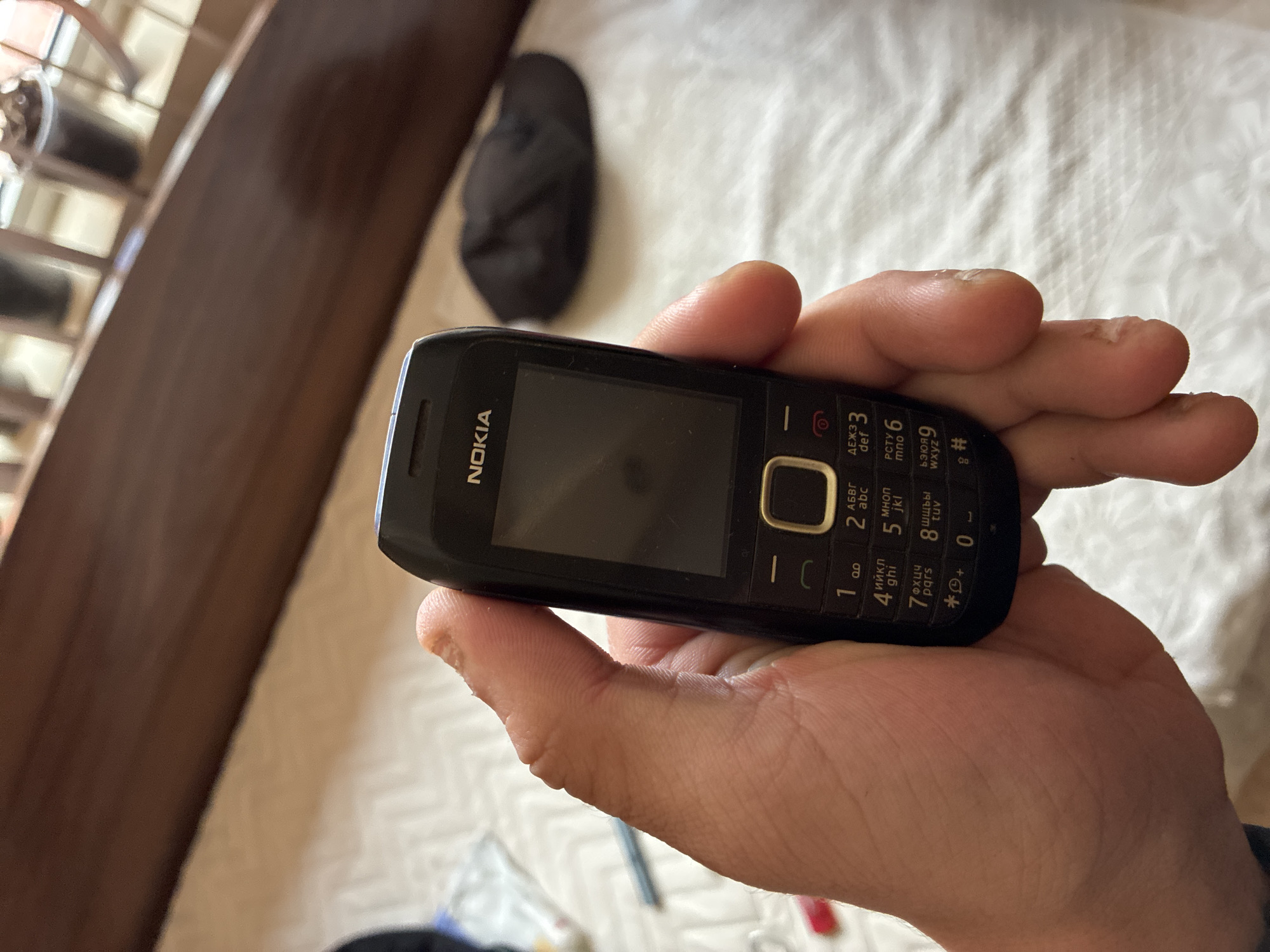 The image captures a close-up of a person's hand holding a black Nokia feature phone. The phone, held diagonally across the frame, features a physical keypad and a dark screen. The brand name "NOKIA" is visible above the screen. The keypad clearly displays numbers 0 through 9, each associated with both Latin and Cyrillic alphabet letters, such as "АБВГ abc" for '2', "ДЕЖЗ def" for '3', "ийкл ghi" for '4', "мноп jkl" for '5', "РСТУ mno" for '6', "фхцч pqrs" for '7', "шщы tuv" for '8', and "ьзюя wxyz" for '9'. Special characters like '*', '#', '+', and '-' are also present.
The background is a soft, light-colored textured fabric, likely a bedsheet or blanket, which is out of focus. A dark, soft object, possibly a slipper, rests on this fabric behind the hand. To the left, a blurry dark wooden structure, possibly a bed frame or a window/balcony railing, partially obstructs the view of what appear to be small potted plants or decorative items. The lighting suggests an indoor setting during daylight. The scene conveys a relaxed, domestic atmosphere, with the hand seemingly holding the phone idly. Based on the provided context, the location is Varna, Bulgaria.