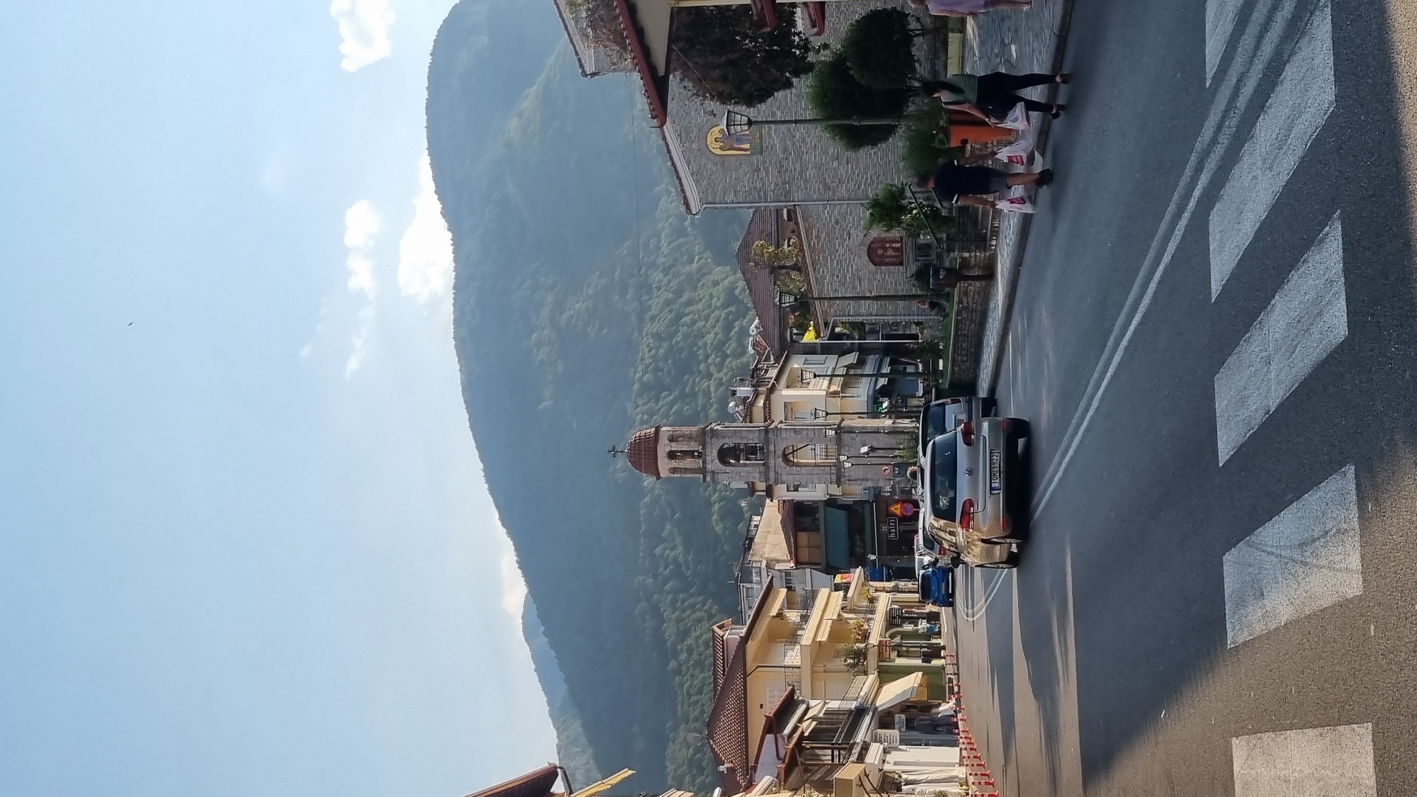 In Litochoro, Greece, a sunlit street in a town is captured. Buildings with red-tiled roofs line the street, with a prominent church tower featuring a red dome rising in the background. The tower has an arched opening with a bell and a small balcony. A gentle slope ascends towards a verdant, tree-covered mountain range that dominates the skyline under a clear blue sky with scattered clouds. Traffic moves along the asphalt road, marked with white lines and a pedestrian crossing in the foreground. A silver Volkswagen Golf is visible driving away, its license plate reading "IOH 3822." Two individuals, one carrying white shopping bags, walk on the sidewalk to the right of the street. The scene is bathed in warm sunlight, casting shadows across the road and sidewalks, indicating it is likely late afternoon.