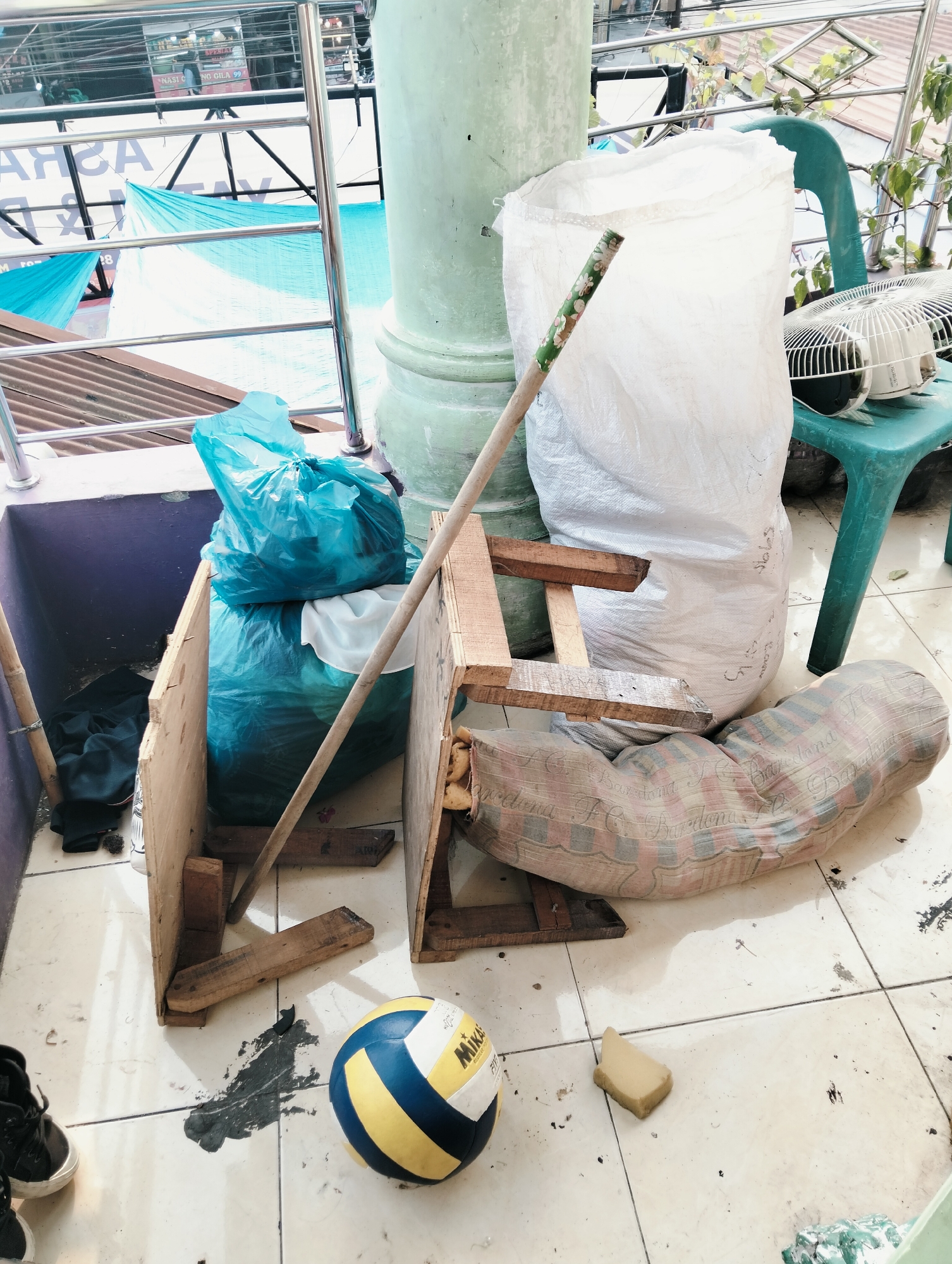 The image depicts an outdoor storage area on a tiled floor, likely in a residential or commercial building in the City of Medan, Indonesia. A turquoise plastic bag filled with items and a white woven bag are prominent, along with a broken wooden chair. A long wooden stick with a decorative handle leans against the chair, and a volleyball lies on the floor nearby. A small fan is placed on a turquoise stool in the background. The scene is cluttered, with various debris scattered across the tiles. The overall impression is one of disarray and neglect.