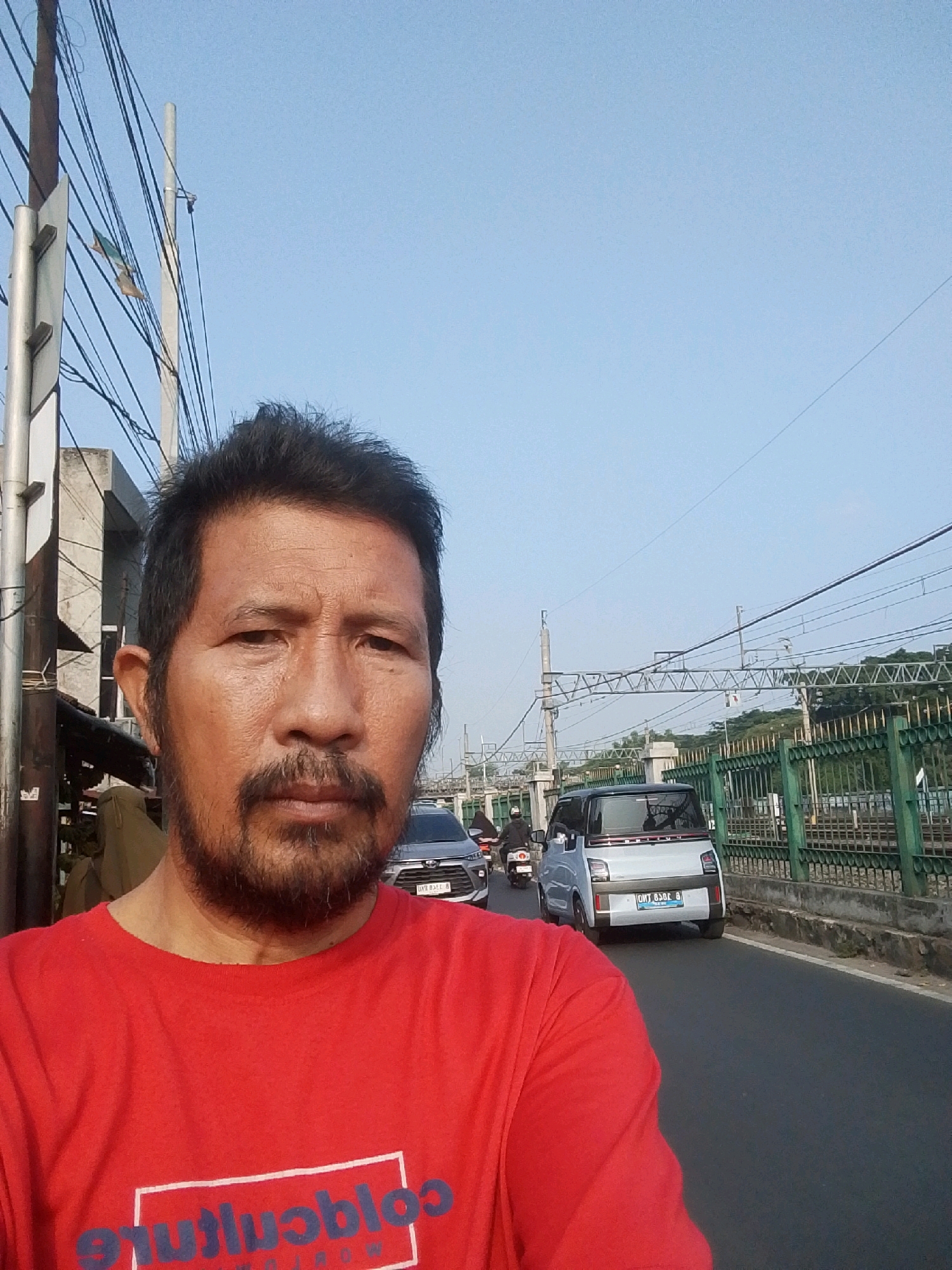 A medium shot selfie captures a man in a red t-shirt standing on a street in East Jakarta, Indonesia. He has a beard and mustache, and appears to be looking directly at the camera. The background shows a street with vehicles, including a white electric car and a motorcycle, and overhead power lines and train tracks. The man is standing near a utility pole and a building. The sky is clear and blue.