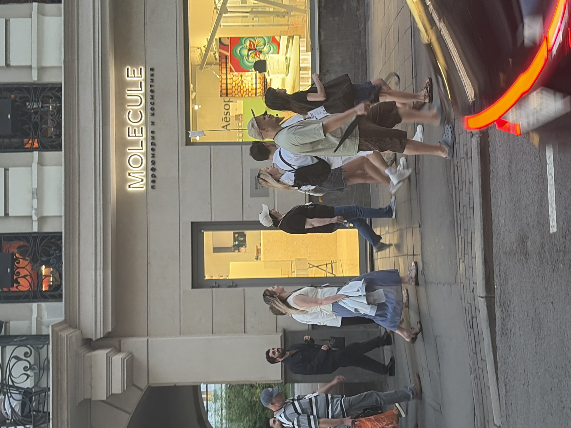 The image shows a street scene in front of a store named "MOLECULE" with Cyrillic text below it, which translates to "perfumery and cosmetics." Several people are walking on the sidewalk in front of the store. The people are dressed in casual clothes and appear to be window-shopping or passing by. A car's rear lights are visible on the right side of the image, suggesting it's moving along the street. The building housing the store has a classic architectural style with decorative details. The scene appears to be set in an urban environment, possibly a shopping district in a city.