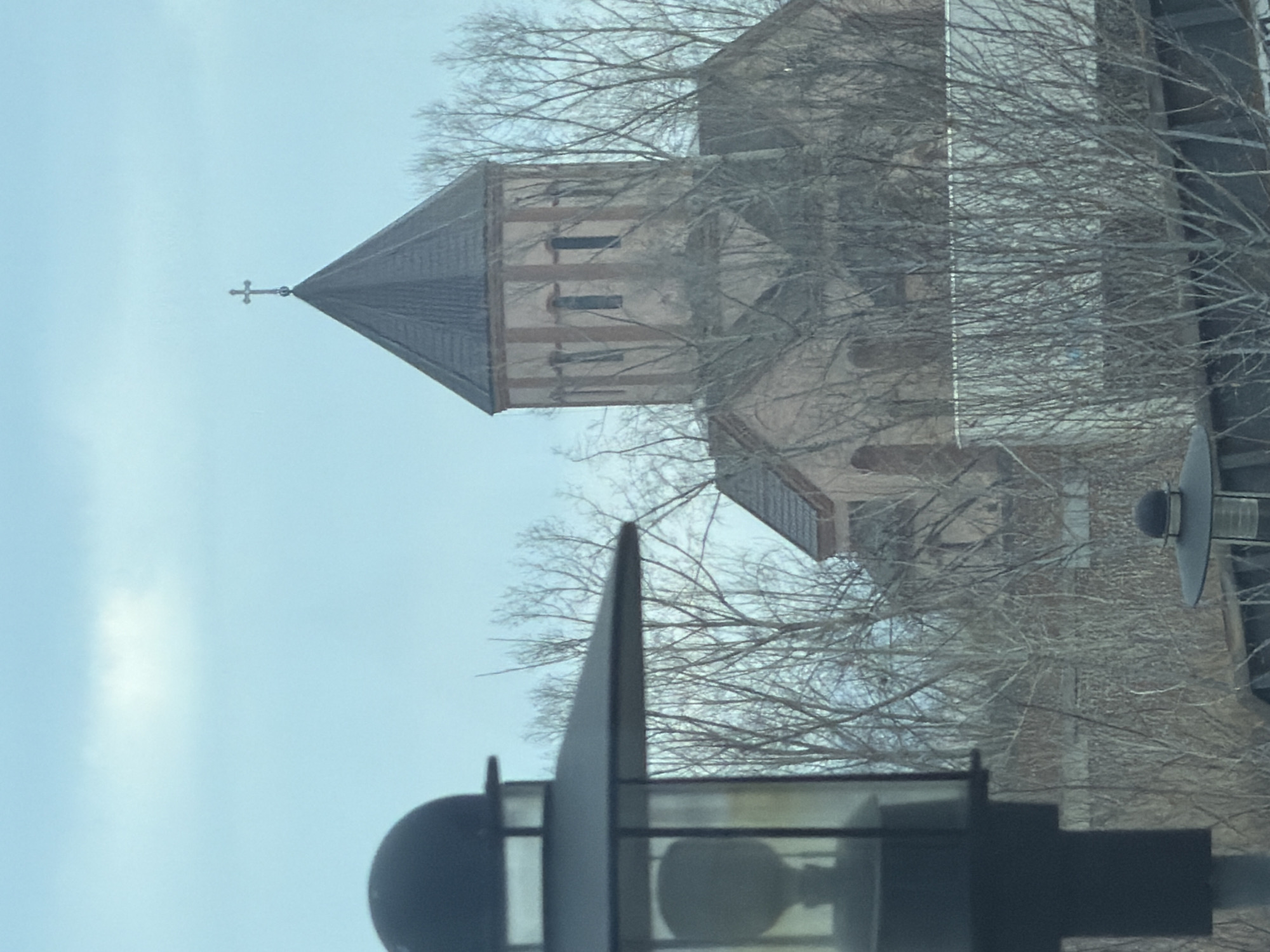The image shows a church steeple with a cross on top, partially obscured by bare trees. In the foreground, there's a street light with a bulb visible inside a glass enclosure, further obscuring the view. The building's architecture suggests it might be located in Krasnoyarsk, Russia, but precise verification of the location requires additional information or context. The sky is a pale blue, indicating either early morning or late afternoon lighting. The overall environment seems urban, though specific details are not readily visible due to the obscuring trees and street light.