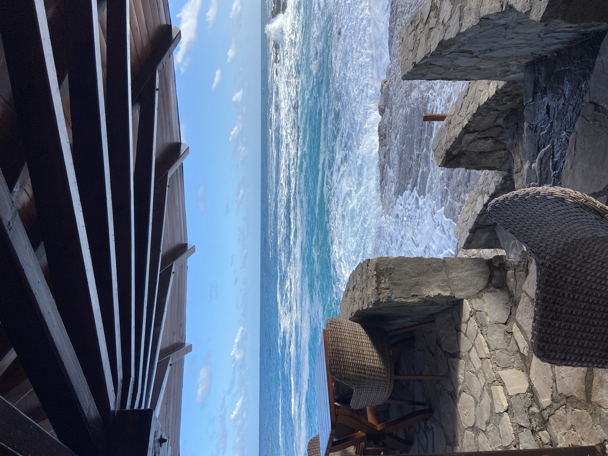 The image shows an outdoor dining area overlooking the Adriatic Sea in Prijevor, Montenegro. The dining area is constructed on a stone terrace with wicker chairs and wooden tables. The sea is visible beyond the terrace, with waves crashing against the rocky coastline. A wooden structure is on the left. The sky is blue with scattered clouds.