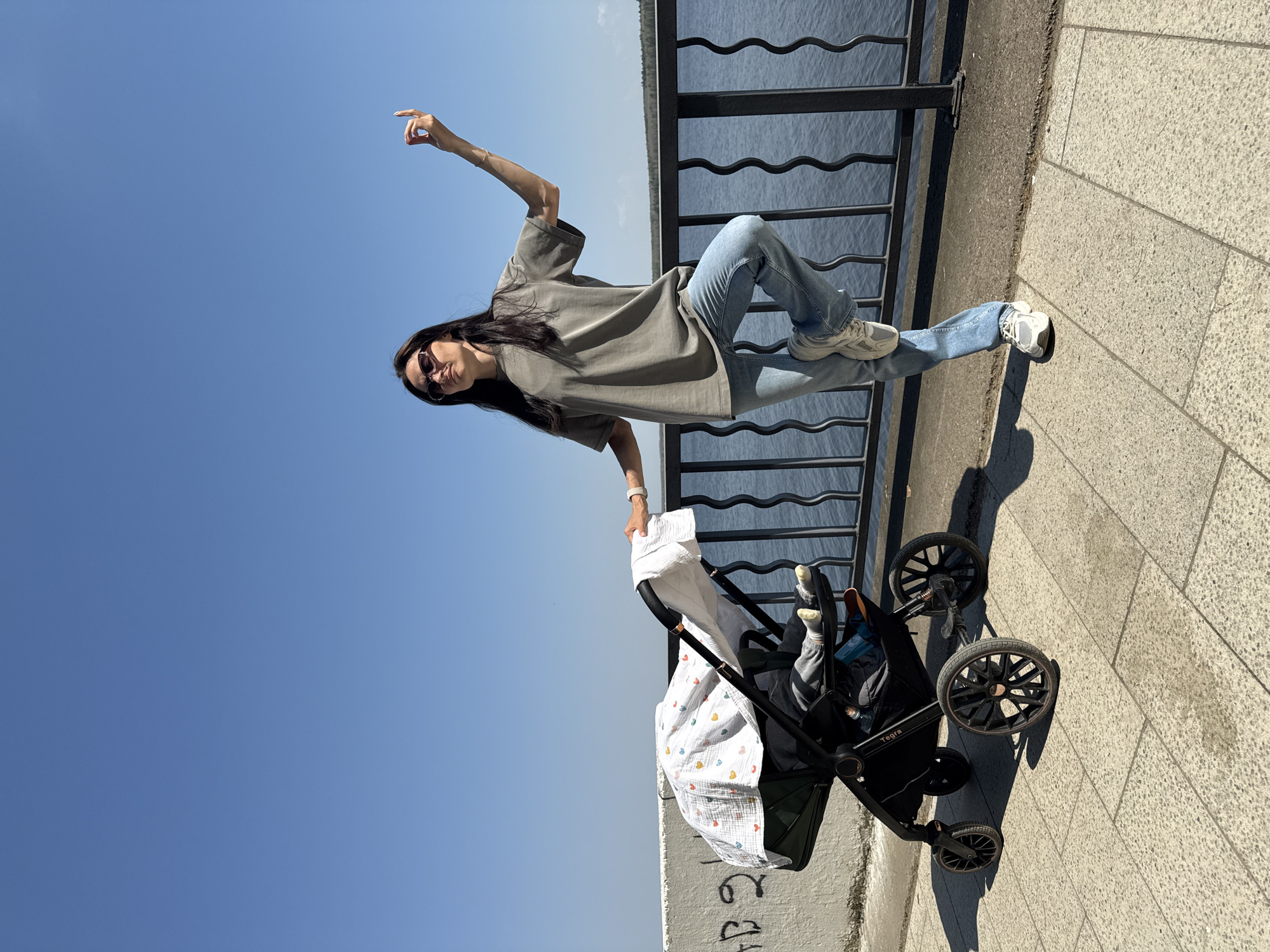 A woman with long dark hair, wearing sunglasses, a gray t-shirt, and jeans is perched on a black metal railing next to a body of water on a sunny day. Her right arm is raised and pointed. She is near a black stroller with a white blanket that has colored hearts. The stroller has a baby inside. The scene appears to be on a paved walkway. Part of Chaikovsky, Russia is visible.