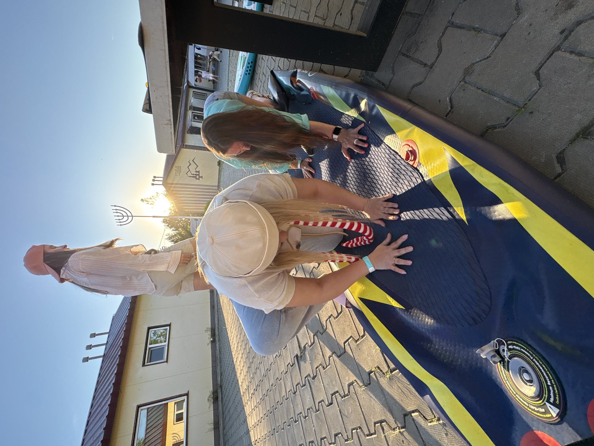 In Birobidzhan, Russia, two women are engaged in an activity on an inflatable paddleboard on a paved area. One woman, wearing a beige cap and a light-colored shirt, is bent over the paddleboard, appearing to adjust or interact with it. A red and white striped lanyard is visible around her neck. The other woman, wearing a teal shirt and shorts, is also on her hands and knees on the paddleboard, facing away from the camera. Her hair is long and brown. A third woman, seen in the background wearing a peach-colored cap and a light-colored shirt, stands facing away from the camera. The scene is illuminated by the setting sun, casting long shadows. In the background, there are buildings with pitched roofs, including one with the text "FOMA" and an image of mountains. A menorah is visible to the left of the frame. The ground is paved with interlocking concrete blocks, with some grass growing between them.