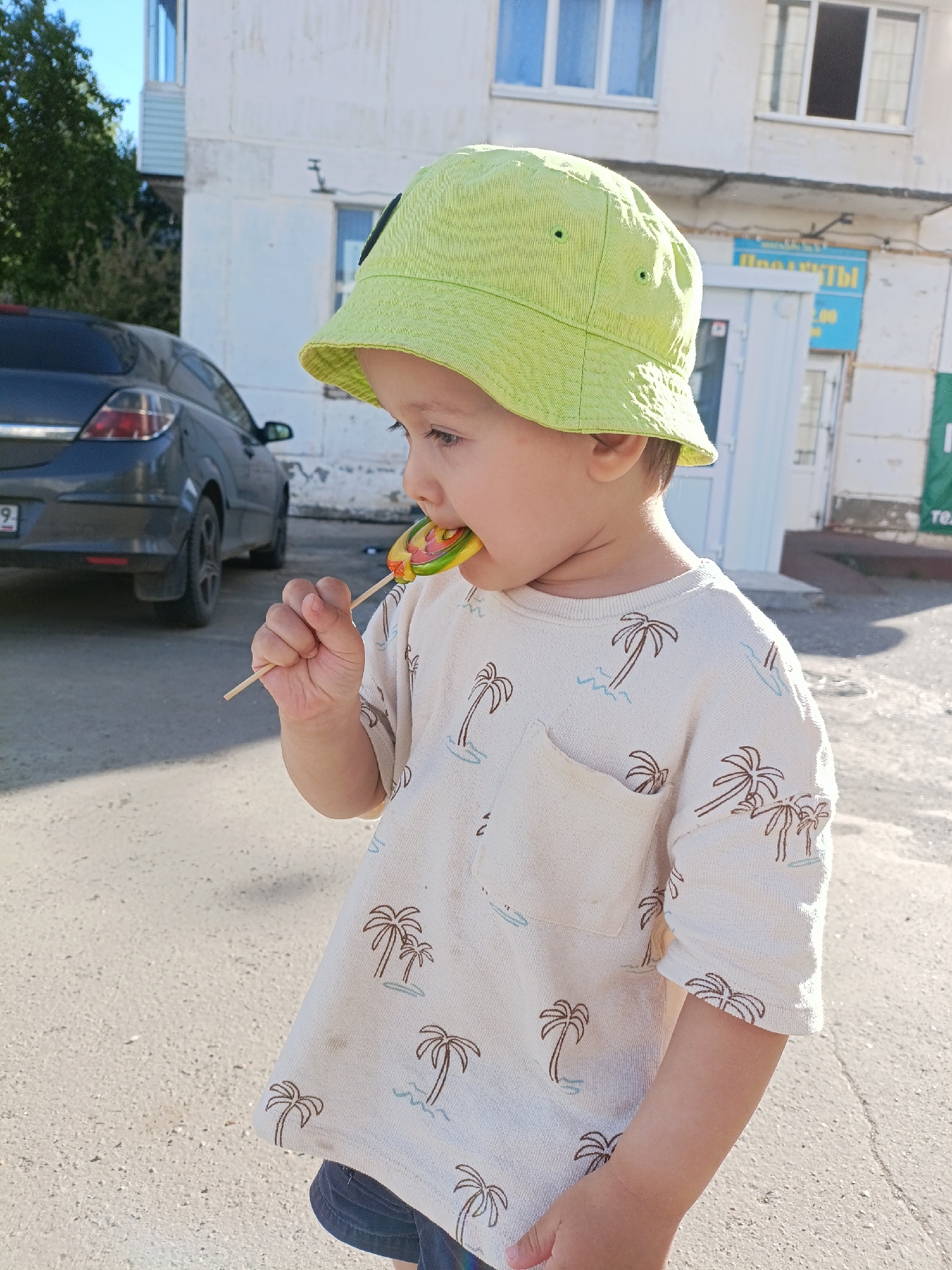 A young child wearing a light green bucket hat and a long-sleeved shirt patterned with palm trees is standing outside with a lollipop in their mouth. They are positioned on a paved surface, possibly a sidewalk or street. In the background, there is a dark gray car and a light-colored building with visible windows and signage in Russian, indicating the location as likely being in the city of Chaikovsky, Russia. The scene is brightly lit, suggesting it is daytime.