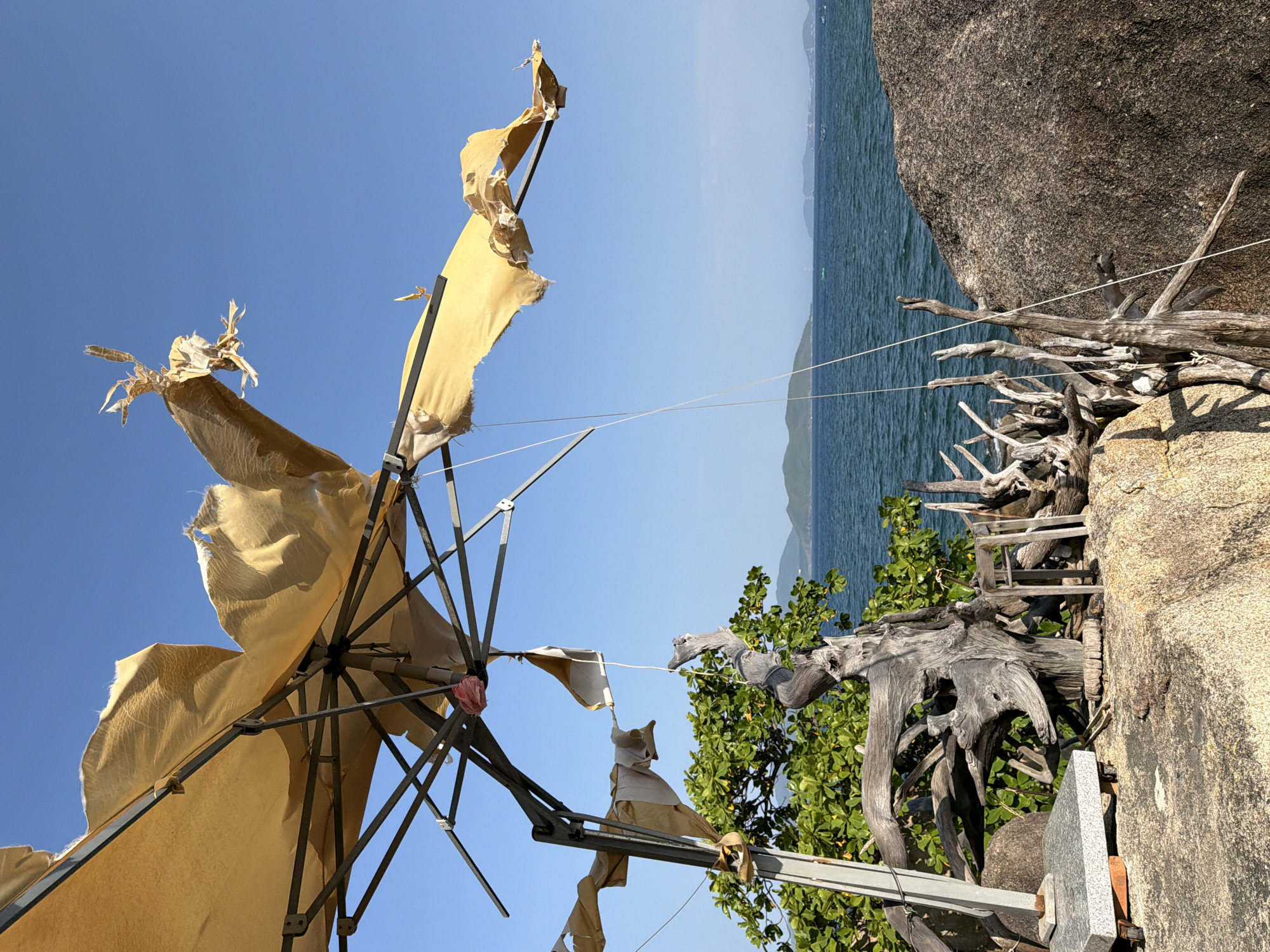This image captures a weathered outdoor scene, likely on a rocky coastal area in Nha Trang, Vietnam, given the presence of the sea, distant islands, and tropical vegetation. A large, tattered yellow umbrella dominates the upper portion of the frame, its fabric ripped and frayed, exposing the metal frame and support structure. White strings hang down from the umbrella, some attached to a piece of driftwood. In the foreground, large rocks form the ground surface, with a collection of bleached and twisted driftwood artfully arranged. A green leafy tree grows behind the driftwood. The sea stretches to the horizon, with faint outlines of islands and what appear to be small boats or buoys visible in the distance. The sky is a clear, vibrant blue.