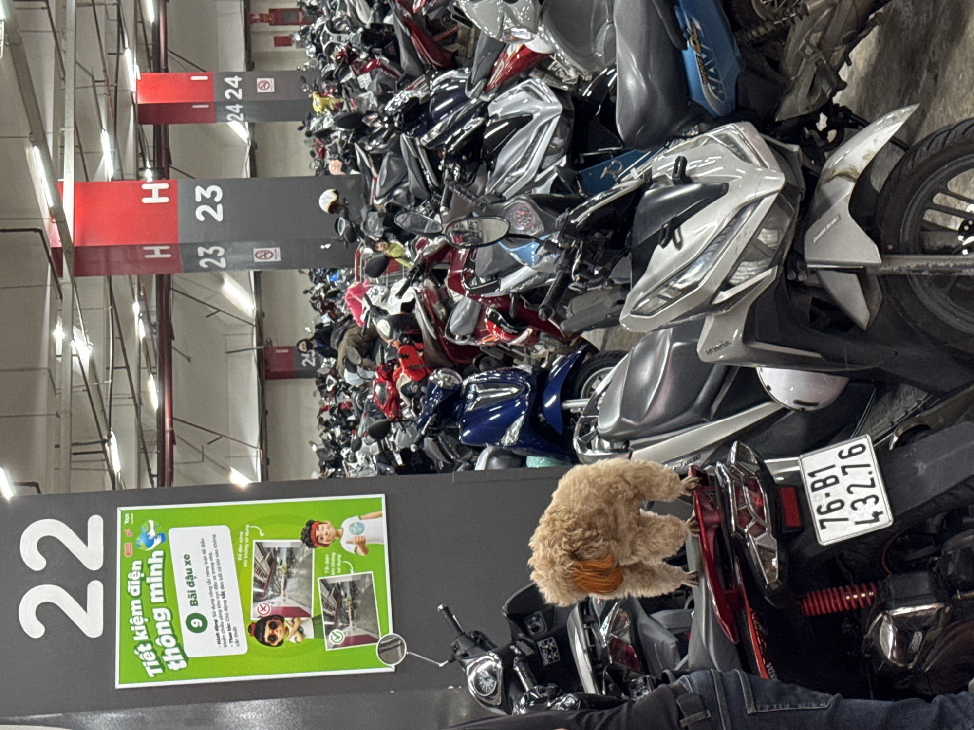 This image captures a dense indoor motorcycle parking garage in what the prompt identifies as the city of Da Lat, Vietnam. In the foreground, a small, light brown, curly-haired dog, possibly a poodle mix with orange-tinted ears, stands on the rear seat of a dark motorcycle. The motorcycle displays a white license plate with black characters "76-B1 432.76". To the left, the lower torso and left leg of a person wearing blue jeans and a dark shirt are partially visible, standing adjacent to the motorcycle the dog is on.
The midground features numerous motorcycles and scooters of various models and colors, tightly packed in rows. A prominent grey vertical column, marked with a large white "22" at the top, has a green information poster affixed to it. The poster's header reads "Tiết kiệm điện thông minh" (Smart electricity saving) and includes a small "Tops" logo. The poster details energy-saving tips with illustrative cartoon figures and images of a parking garage, one showing lights on (with a red 'X') and another with lights off (with a green checkmark).
Further into the background, several additional support columns are visible. These columns are painted in alternating sections of red and dark grey. One column is marked "H 23 23", and another slightly further back shows "I 24 24", indicating parking section identifiers. Another column to the far left has the number "25" partially visible. The ceiling structure reveals exposed utility pipes and fluorescent lighting fixtures, characteristic of a commercial parking facility. The lighting is bright and artificial, casting a uniform illumination over the crowded scene.