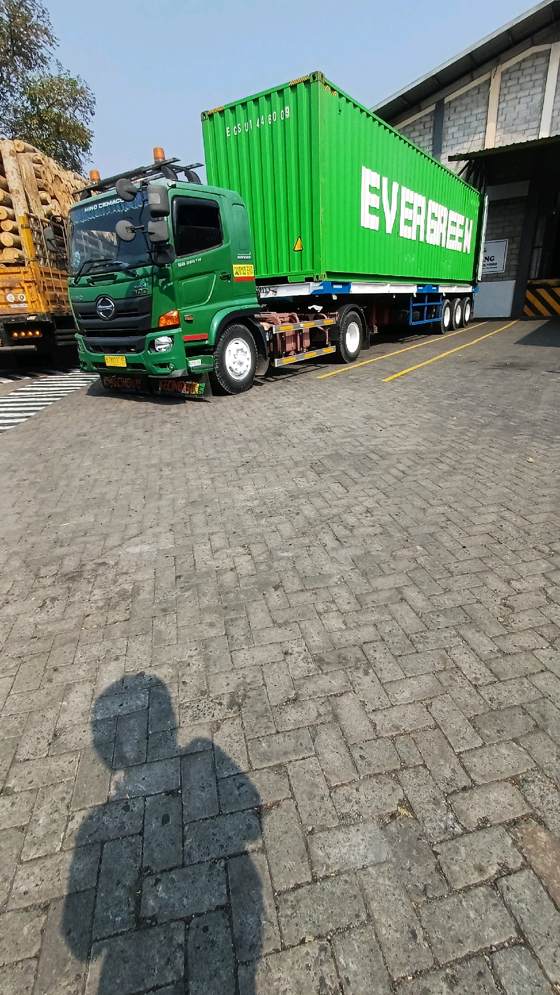 An outdoor, daytime, medium-shot photograph captures a green Hino truck parked on a cobblestone surface in what appears to be a logistics or industrial area. The truck is hauling a large, vibrant green shipping container with "EVERGREEN" prominently displayed in white lettering. The container has identification markings on its side, including "E CS U1 44 60 09".
The truck cab is green, with Hino branding visible on the front grille. It has orange hazard lights on its roof. The trailer supporting the container is blue and has multiple axles with white wheels. To the left of the Hino truck, another truck is partially visible, loaded with logs.
The background features a building with a corrugated metal roof and siding, suggesting it could be a warehouse or factory. Yellow lines are painted on the ground, indicating parking or traffic lanes. A shadow of a person, presumably the photographer, is cast on the cobblestones in the foreground. The sky is clear and blue, indicating a sunny day. The location is identified as Kranggan, Indonesia.