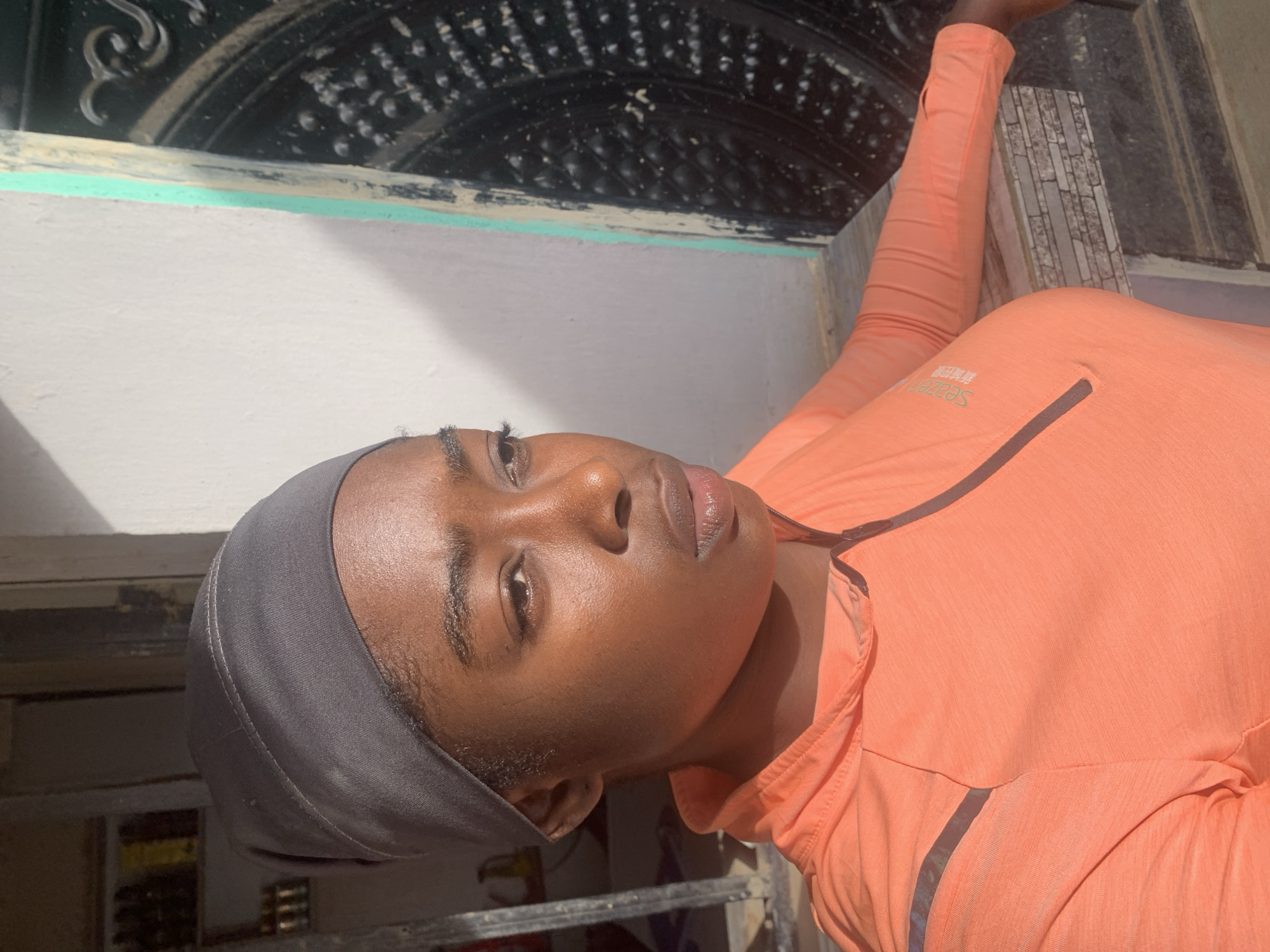 A person is posing indoors, possibly near a window or doorway. The individual is wearing a gray head covering and a light orange, long-sleeved shirt. The background features a textured, dark, possibly metallic or intricately designed structure, with hints of light blue, white, and gray surrounding it. A tiled wall is visible in the lower right portion of the image. The location might be in the vicinity of New Stadium, Nigeria.
