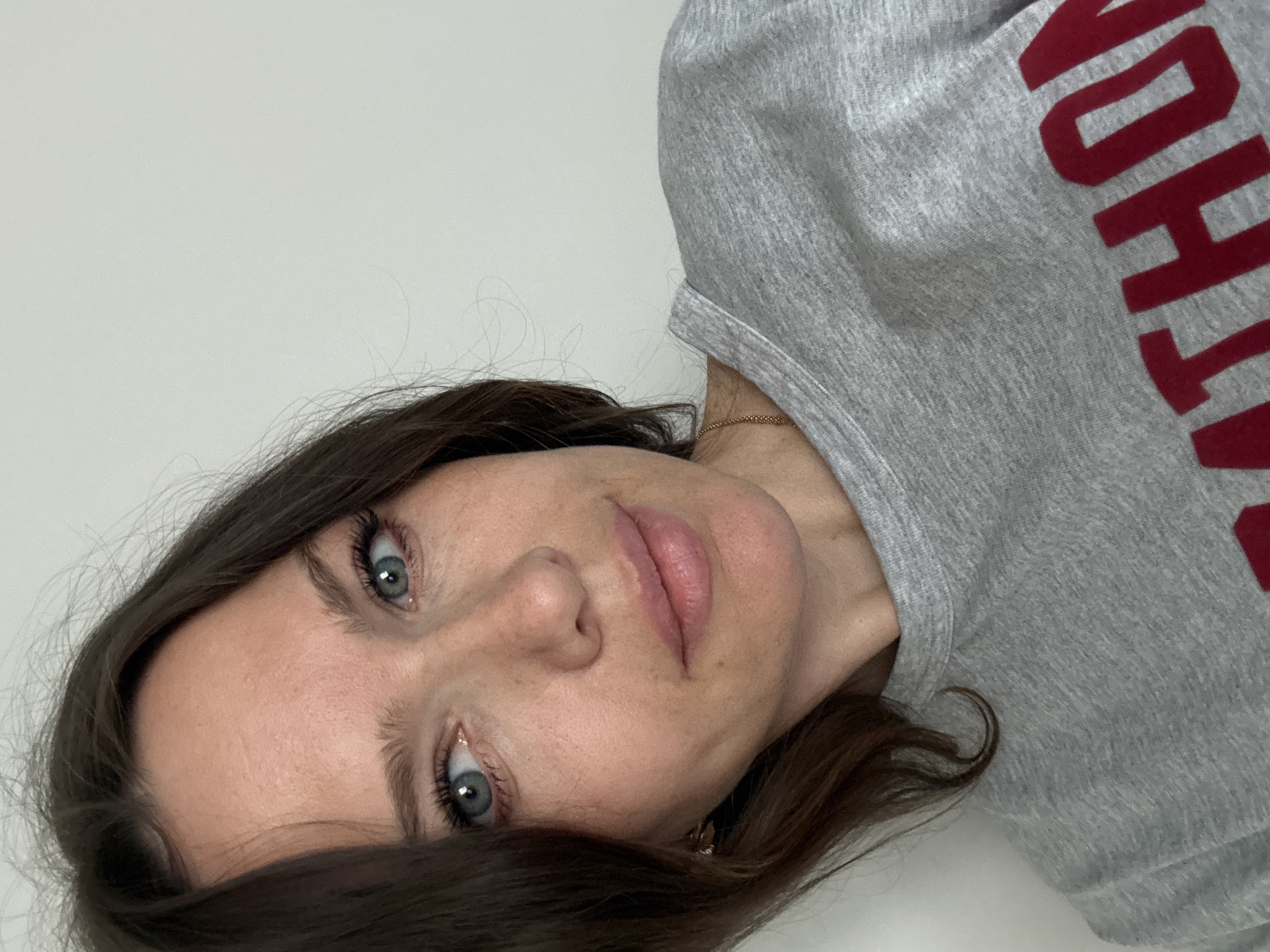 A woman is pictured in a close-up selfie. She has fair skin, dark brown hair, and blue eyes. She is wearing a gray sweatshirt with red lettering that appears to say "THON." A gold chain necklace is visible at her neck. The background is a plain white wall. The setting is Unknown, Unknown.