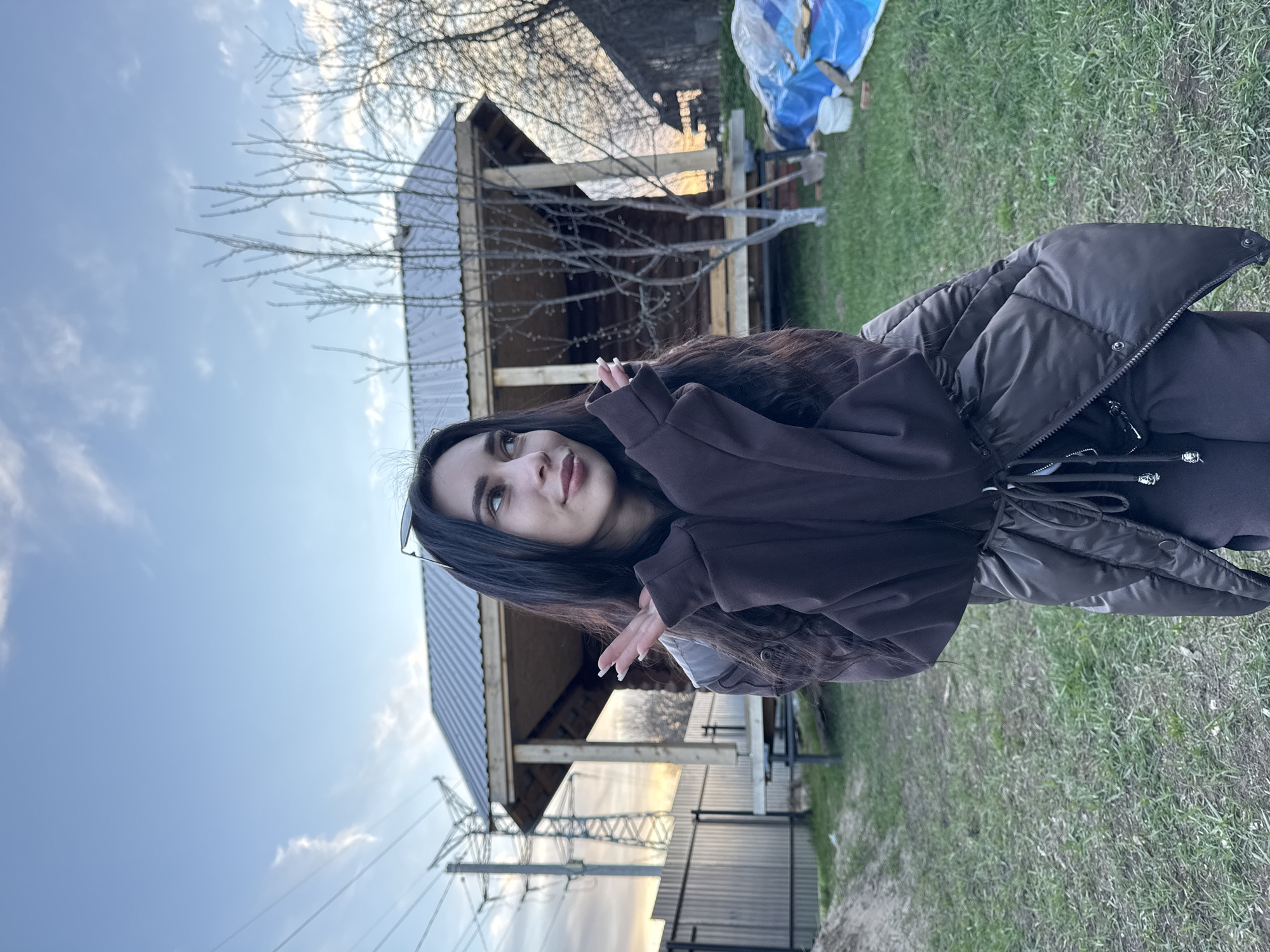 The image shows a woman in a brown hoodie and black puffer jacket, standing outdoors. Behind her is a wooden structure with a metal roof under construction. There's green grass and a tree without leaves in the background. Power lines and a cloudy sky are visible in the distance. The location is Chaikovsky, Russia.