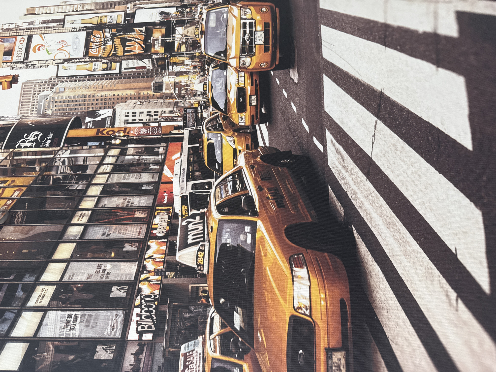 The image depicts a bustling urban street scene, characterized by numerous yellow taxis and towering buildings adorned with bright advertisements. The architectural style suggests a major metropolitan city, likely New York City's Times Square, given the presence of iconic yellow cabs and the vibrant, overwhelming display of digital billboards. The time of day appears to be daytime, with the sunlight casting shadows on the zebra crossing in the foreground, creating a dynamic visual effect. The scene is filled with motion, with cars and possibly pedestrians implied by the activity, conveying a sense of energetic urban life. The image does not depict the city of Anudinovka, Russia.