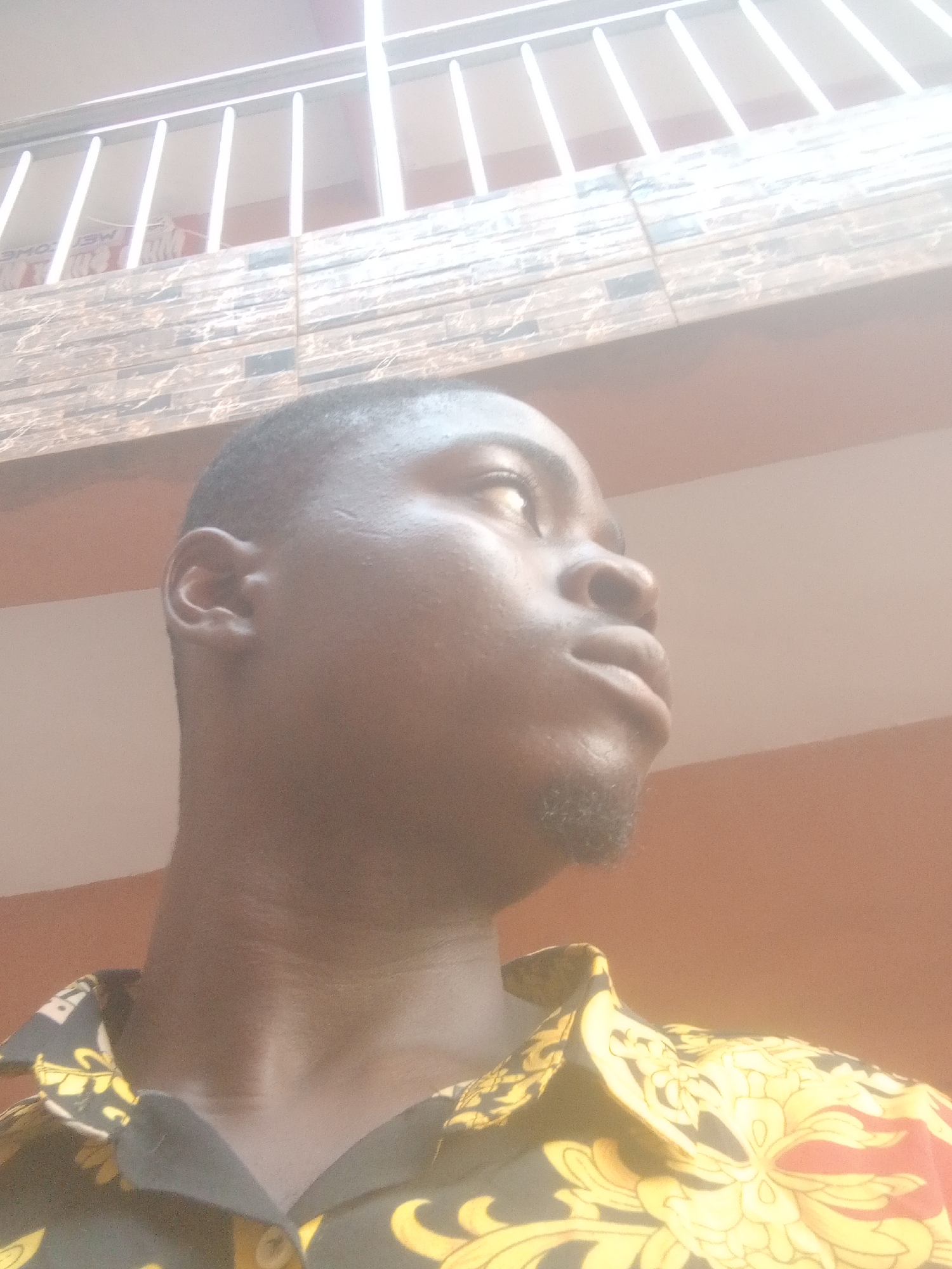 A medium shot looking upward shows a man with a dark complexion. He has short, dark hair and a trimmed beard. He is wearing a patterned shirt with yellow, black, and red designs. Above him is a light-colored wall with a patterned ledge. A white railing is visible above the ledge. The picture seems to be taken outside in Nkpor, Nigeria.