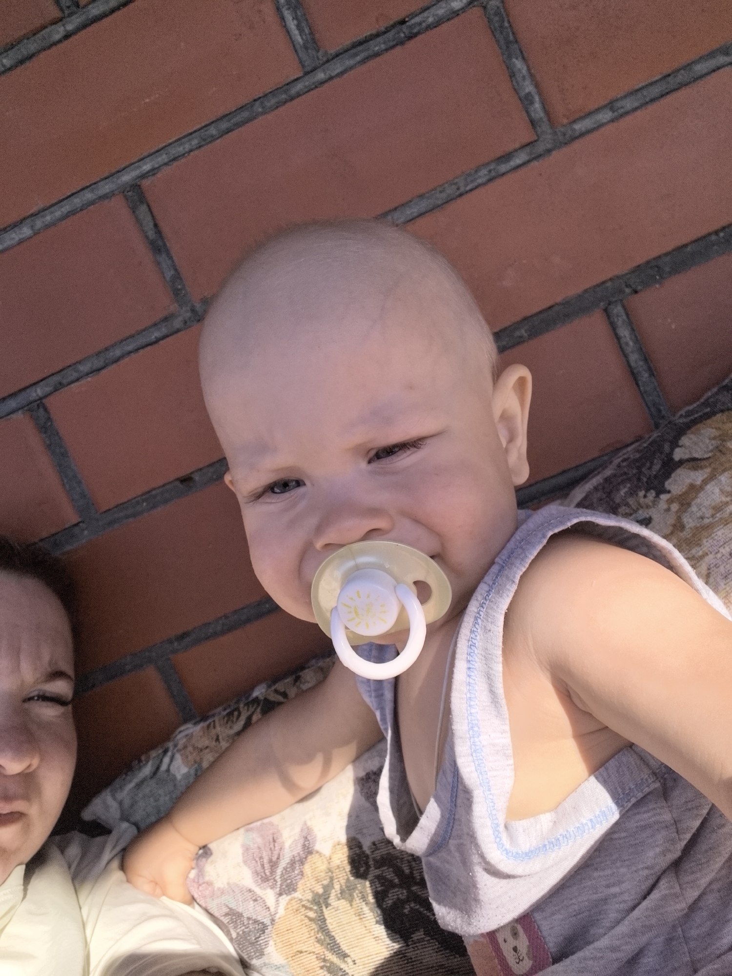 A close-up, low-angle selfie shows a young child and an adult lying down. The child, in the foreground, has fair skin, light-colored eyes, and is wearing a gray tank top. The child has a pacifier in their mouth. The adult, partially visible in the lower left corner, has darker hair and is looking at the camera with a strained expression. They are both lying on what appears to be a cushioned surface with a floral pattern. In the background, a red brick wall is visible. The lighting suggests an outdoor setting, possibly during the day. The image appears to have been taken in Vityazevo, Russia, given the presence of a red brick wall commonly found in the area.