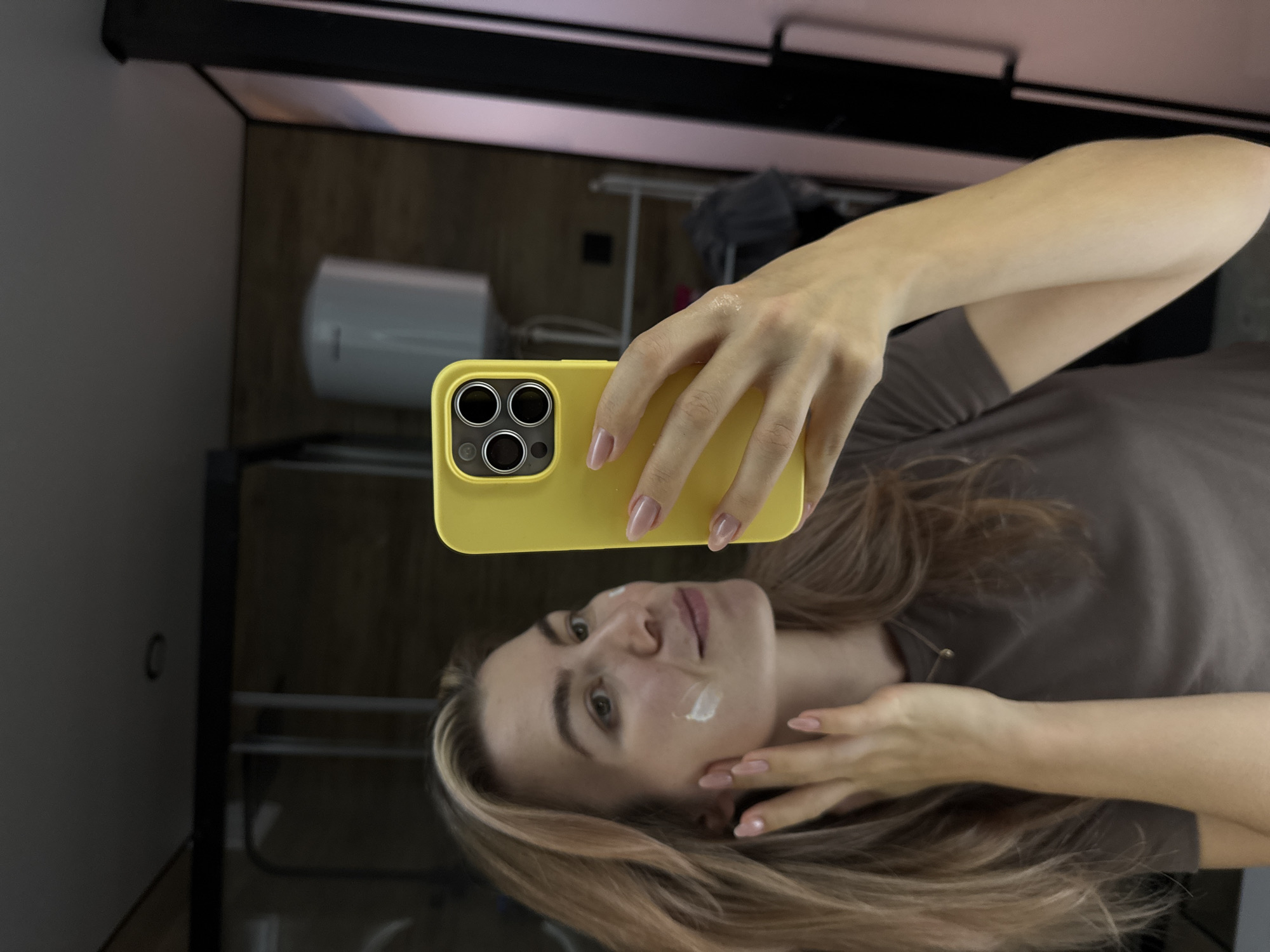 A selfie shows a woman holding a yellow cased smartphone. She has blonde hair and a small amount of white cream on her cheek. Her nails are manicured. She wears a brown shirt. The reflection in the glass behind her shows a white water heater mounted on a dark wall. Unknown, Unknown.