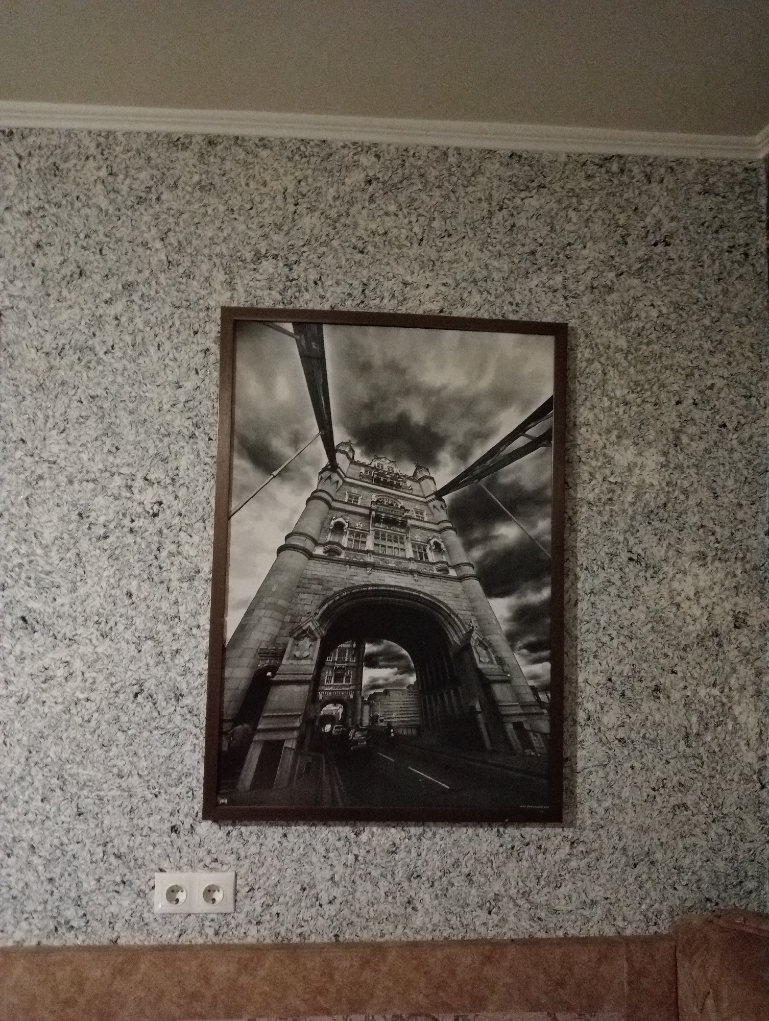 A framed black and white photograph of the Tower Bridge in London is displayed on a wall with textured, grey wallpaper. The photograph, viewed from a low angle looking upwards, features the bridge's towers and the roadway passing underneath. In the lower left portion of the image, a white electrical outlet with two circular sockets is visible on the wallpapered wall. The lower section of the wall transitions to a different textured material, possibly upholstery.