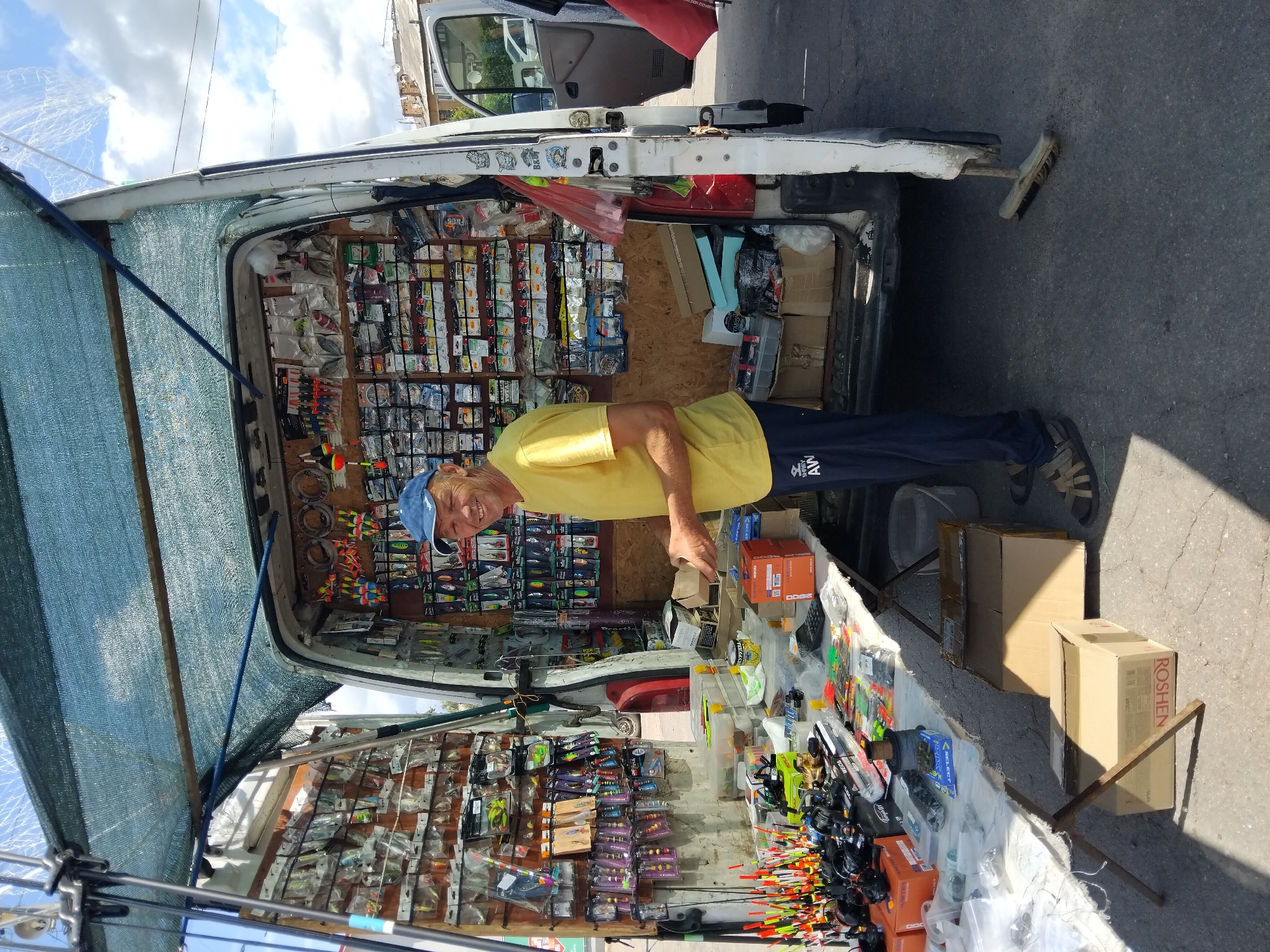 A man in a yellow t-shirt and a blue baseball cap is standing in front of an open-backed white van, which is acting as a stall selling fishing gear. The van's interior is packed with fishing lures, floats, lines, and other accessories, organized on shelves and pegboards. The man is holding a small box and appears to be looking at the camera with a smile. In front of the van, a table displays more fishing equipment, including fishing rods and reels. The scene is outdoors, with a street or paved area in the foreground and buildings visible in the background, suggesting an urban or semi-urban setting. The sky is partially cloudy. The overall impression is a market stall or roadside vendor.