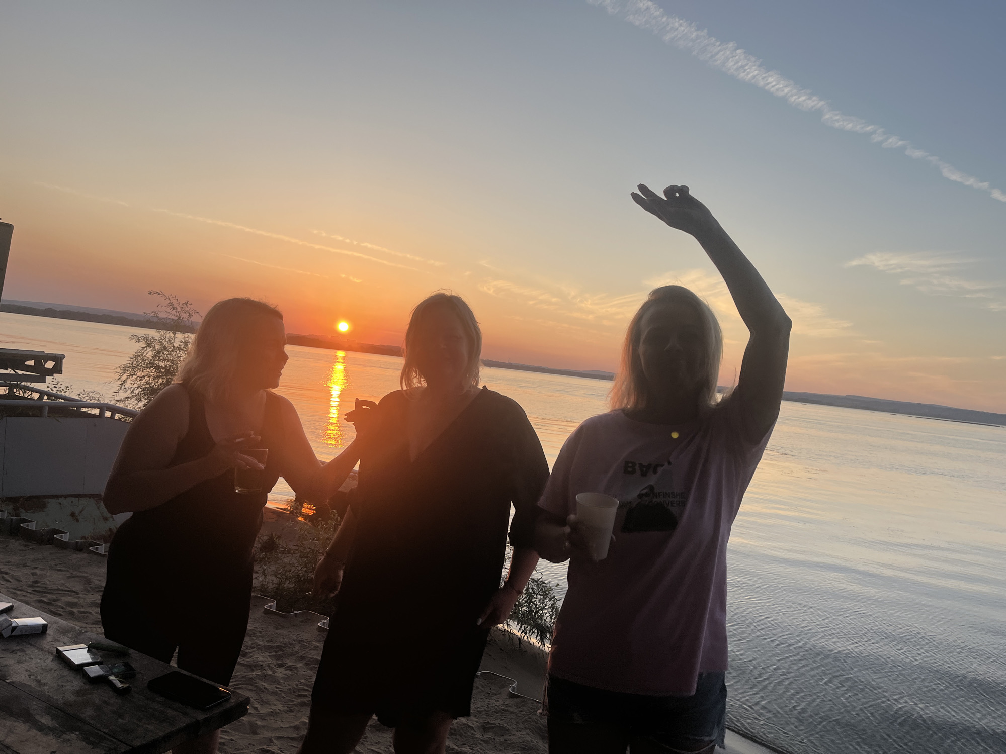 Three women are standing on a sandy beach during sunset. The sun is visible on the horizon over a body of water. The woman on the left is wearing a black dress and holding a drink. The woman in the middle is wearing a black dress. The woman on the right is wearing a pink t-shirt and denim shorts and has her arm raised. There is a table in the foreground with cigarettes and a phone on it. The scene is taking place in Shumeika, Russia.