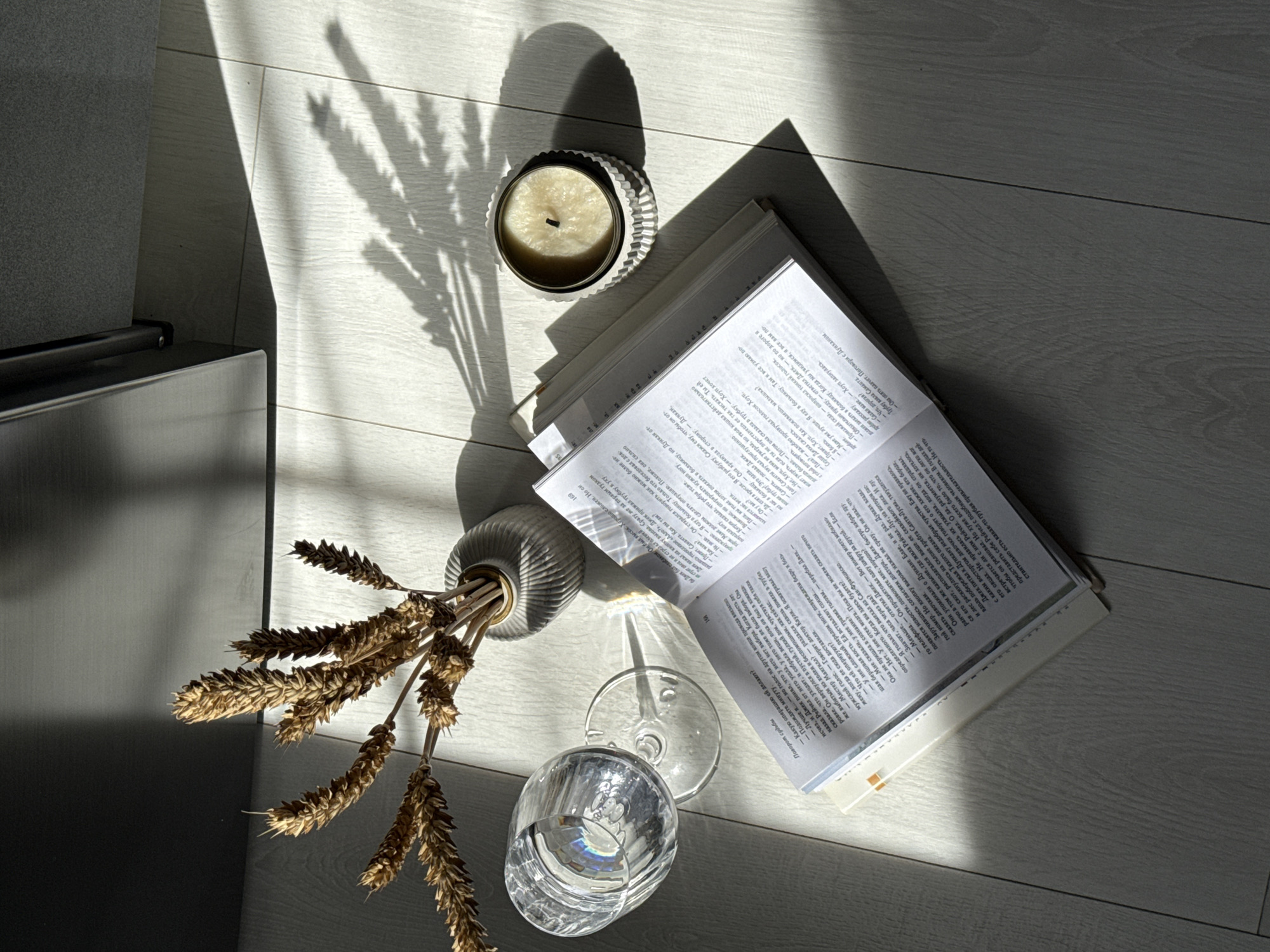 The image shows a still life arrangement on a wooden floor, illuminated by strong sunlight casting shadows. A book is open in the center, its pages filled with text. To the left of the book is a wine glass filled with water, and to the right is a lit candle in a decorative holder. Behind the book, a small vase contains dried wheat stalks. The overall scene is calm and serene, bathed in the warm glow of the sun.