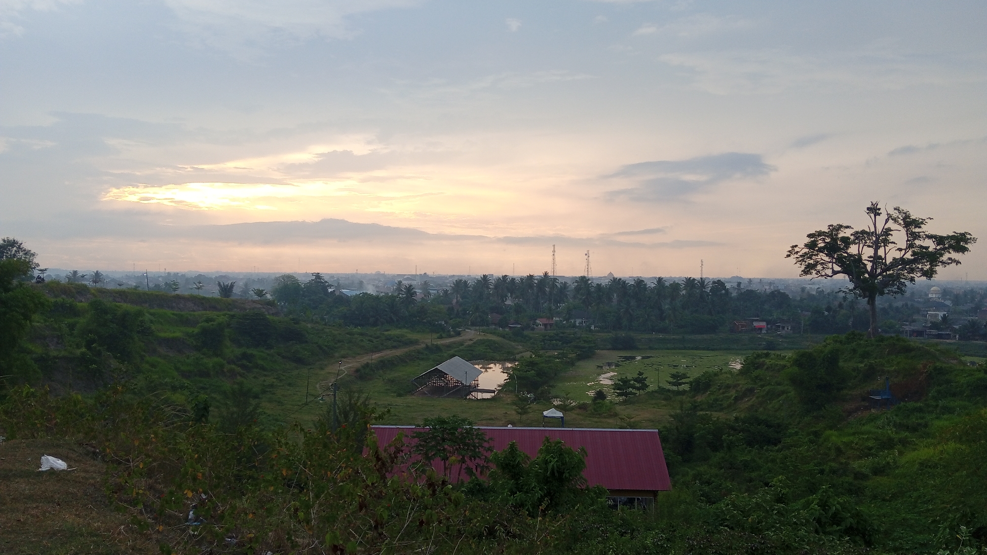 The image depicts a landscape in Uruekanoe, Indonesia, at sunset or sunrise. In the foreground, lush green vegetation dominates with a building having a red roof partially obscured by trees. Further back, a small pond with a structure stands out amidst the greenery. Beyond the pond, a dense line of trees stretches across the midground. In the distance, communication towers and a mosque are visible on the horizon. The sky is filled with soft, muted colors of the setting or rising sun, interspersed with clouds. A large tree stands prominently on a raised area to the right. A patch of waste material is visible on the ground in the lower left.